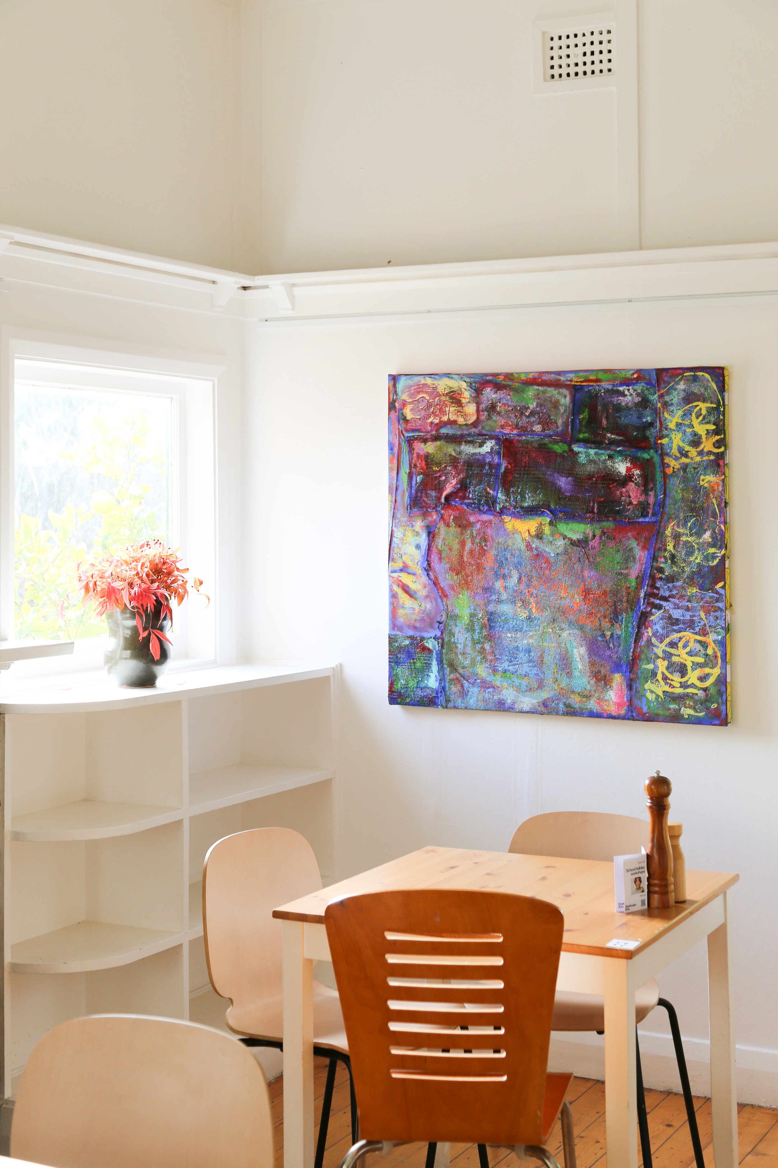 A dining area with a wooden table and beige chairs, a window with a vase of pink flowers, a colorful abstract painting on the white wall, and a white corner shelf.