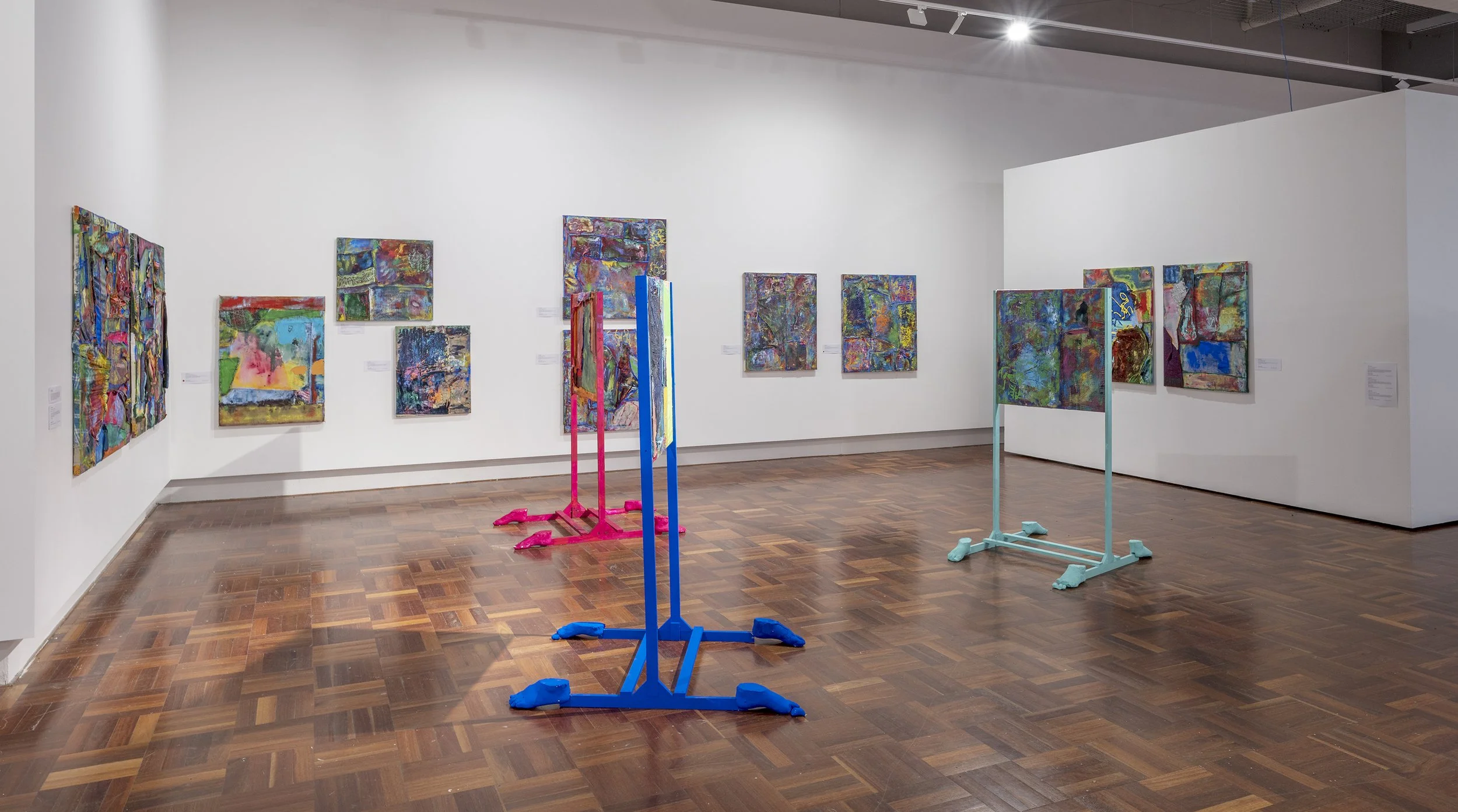 An art gallery with colorful abstract paintings on white walls and three colorful frames on the wooden floor, one pink, one blue, and one light blue.