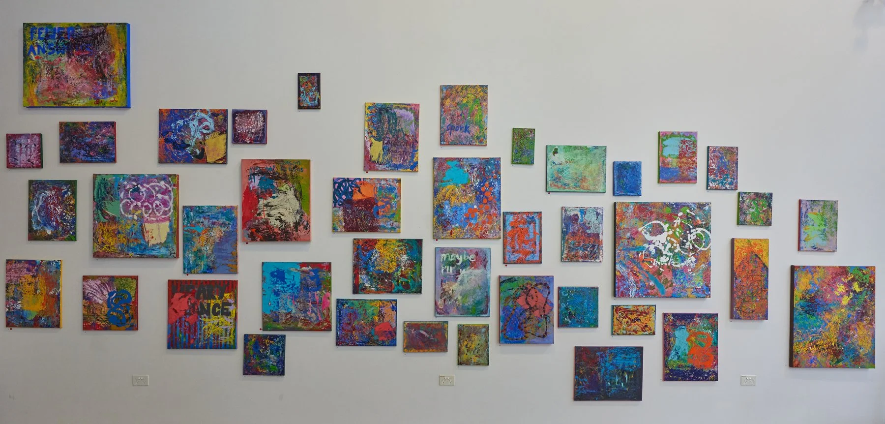 An abstract art gallery wall featuring numerous colorful, textured mixed media paintings varying in size, with some containing text and various vibrant patterns.