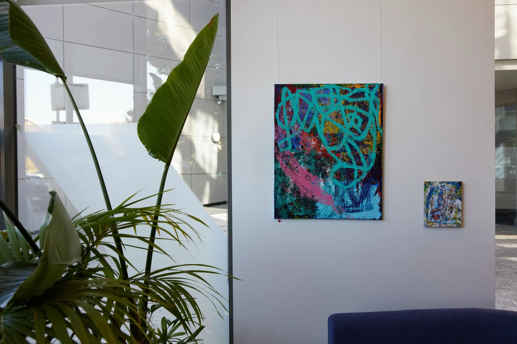 Interior of an art gallery with two abstract paintings on a white wall, a large green potted plant in the foreground, and a blue sofa partially visible at the bottom right.