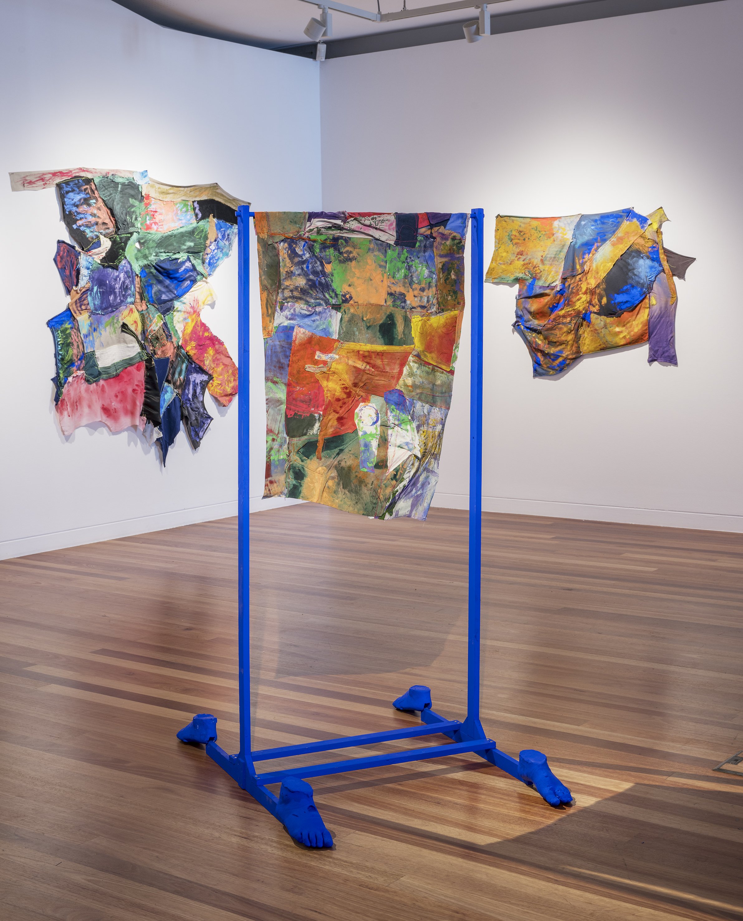 Mixed media textile art installation displayed on a blue frame in an art gallery with wooden floors and white walls.