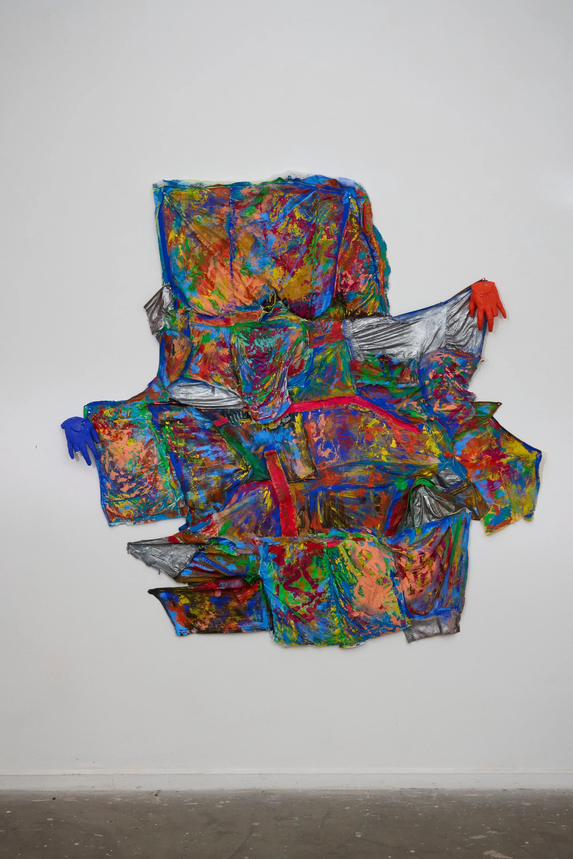 Mixed media artwork resembling a chaotic, colorful sculpture of a person with outstretched arms, wearing orange and blue gloves, against a white gallery wall.