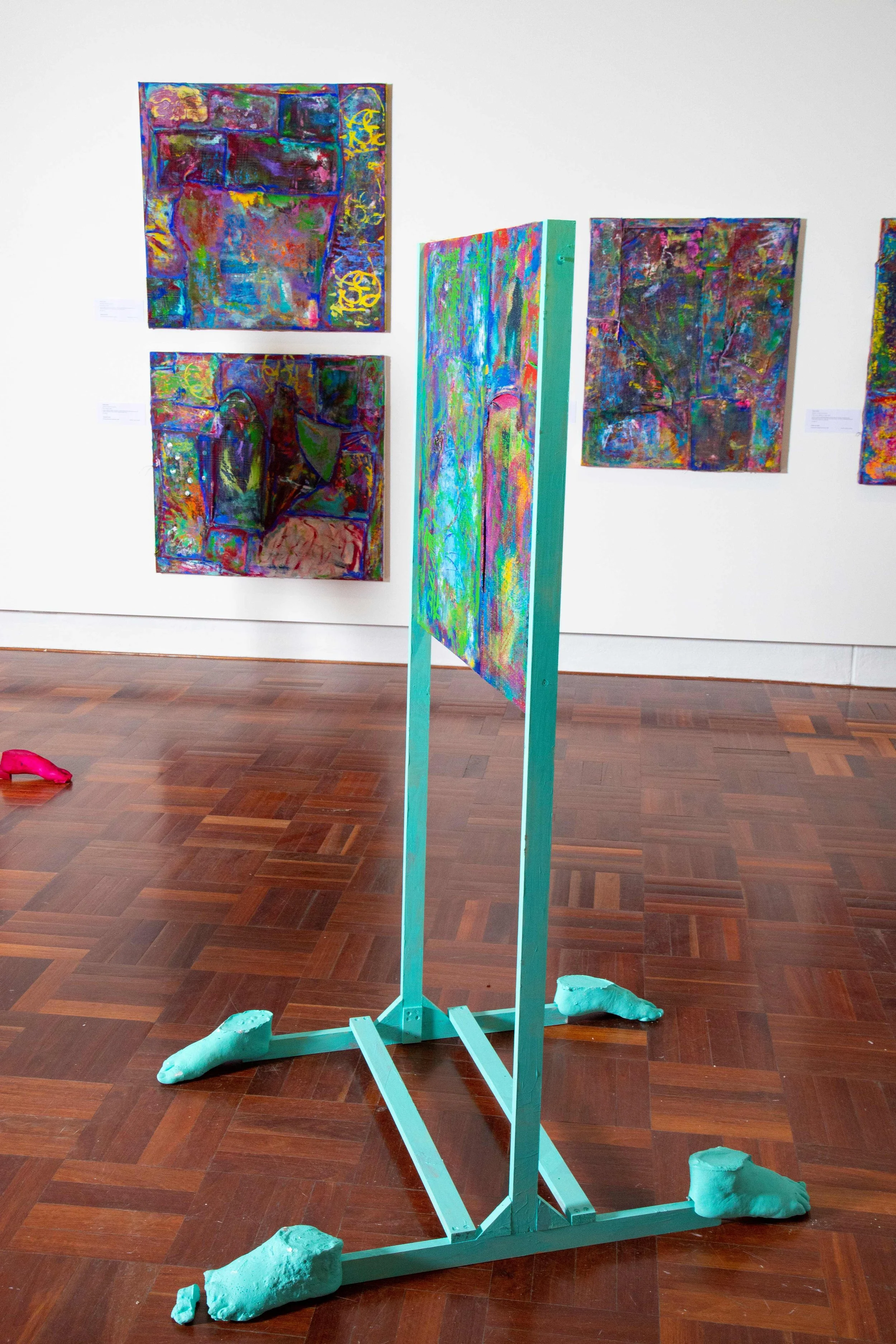 Colorful abstract paintings displayed on a white gallery wall and a turquoise freestanding art piece on the hardwood floor.