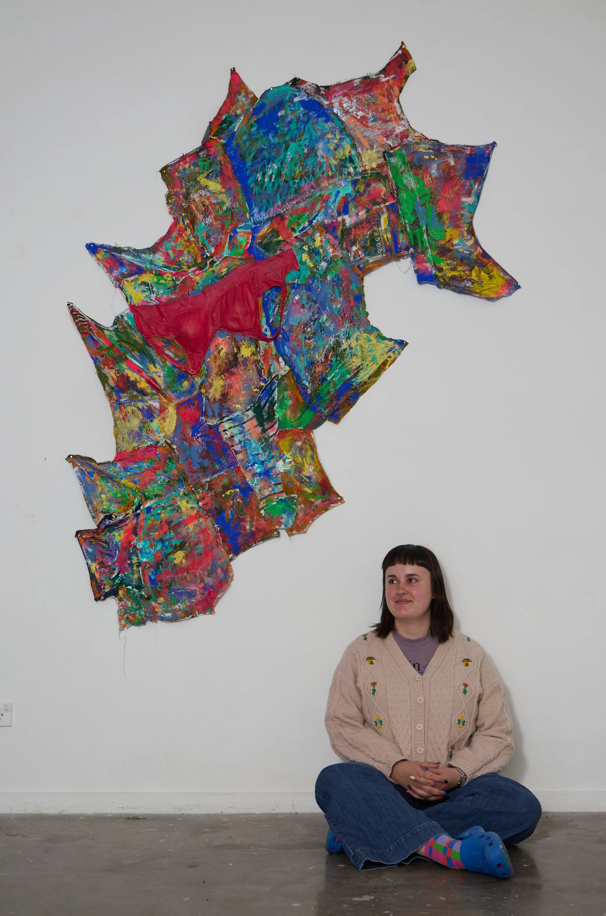 A woman sitting on the floor in front of a white wall with a colorful abstract art piece hanging above her. She has shoulder-length dark hair with bangs and is wearing a tan cardigan with embroidery, blue jeans, and multi-colored socks.