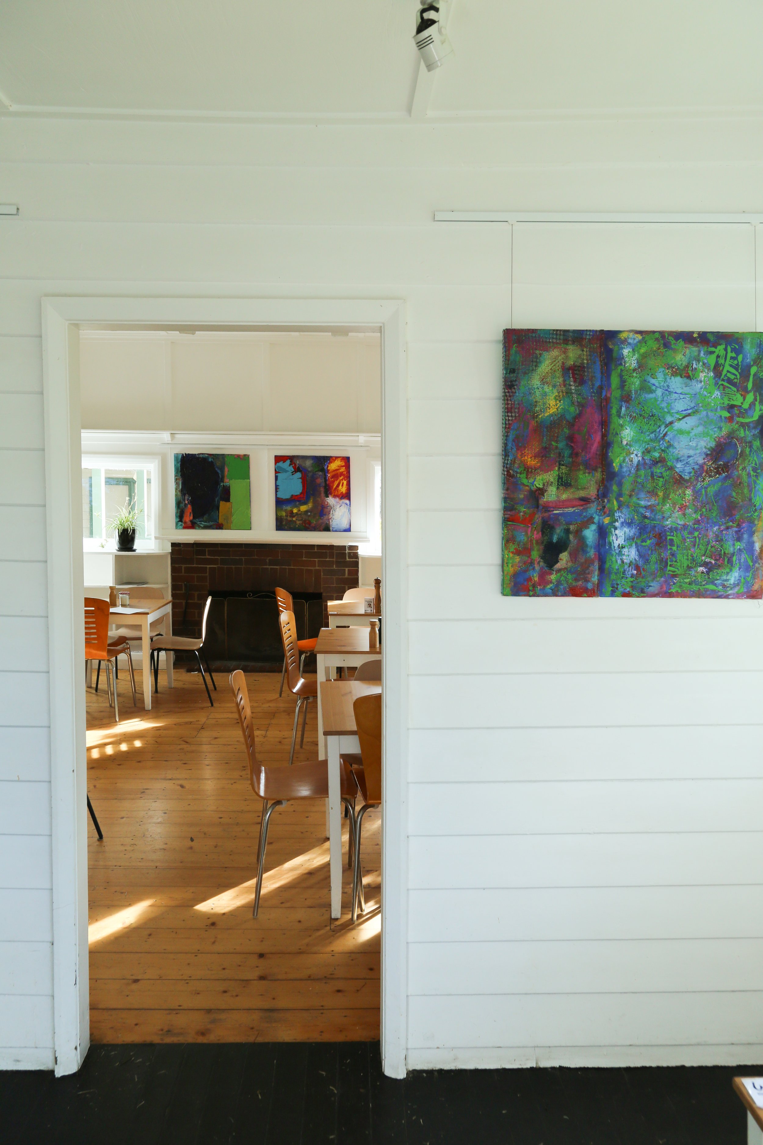 Interior view of a cozy cafe or restaurant with wooden flooring, white painted wood walls, and colorful abstract artwork hanging. The room has wooden tables and chairs, with a brick fireplace visible at the back and a window with a potted plant.
