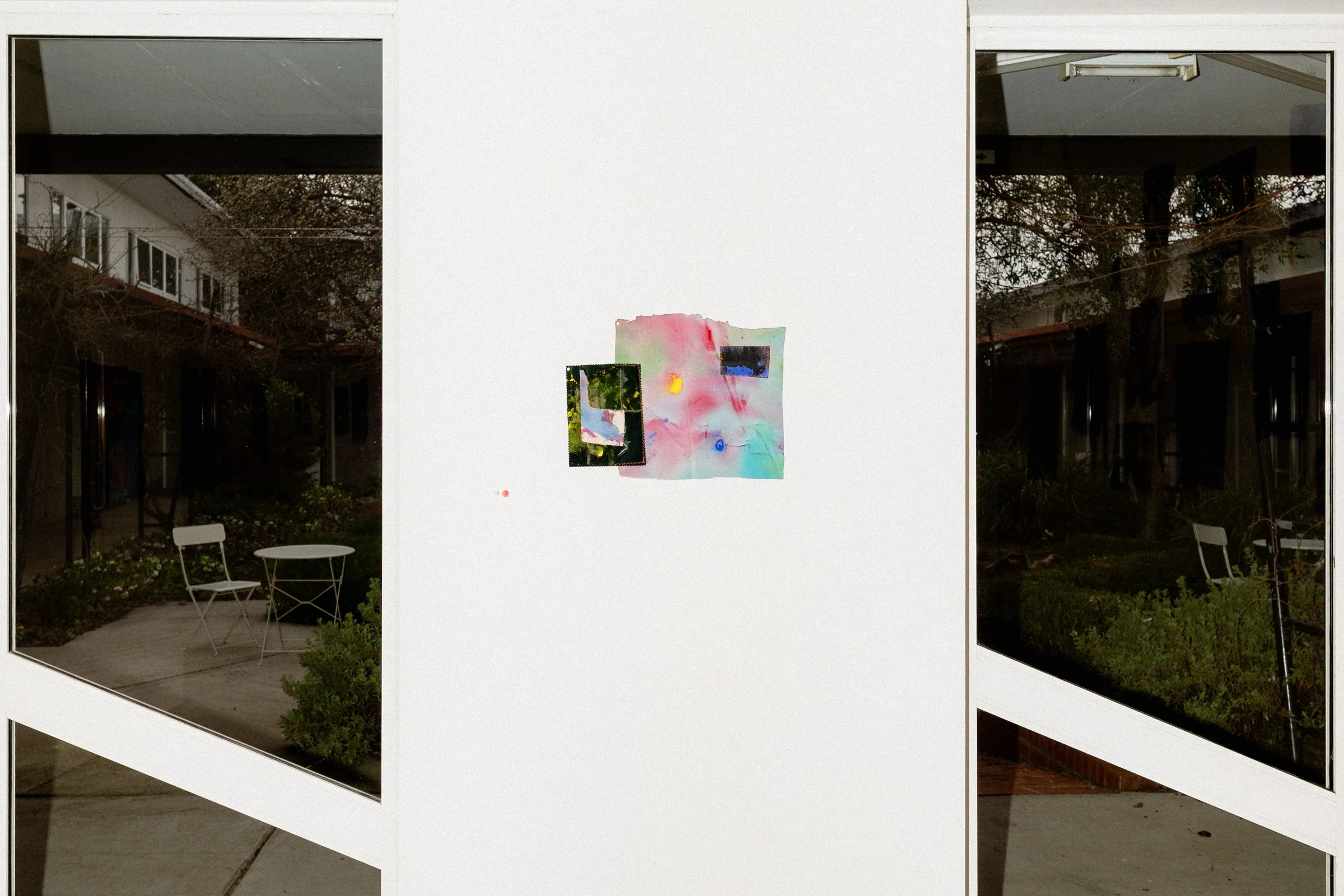Art display with colorful abstract painting on a white wall, reflected in glass windows showing outdoor furniture and garden
