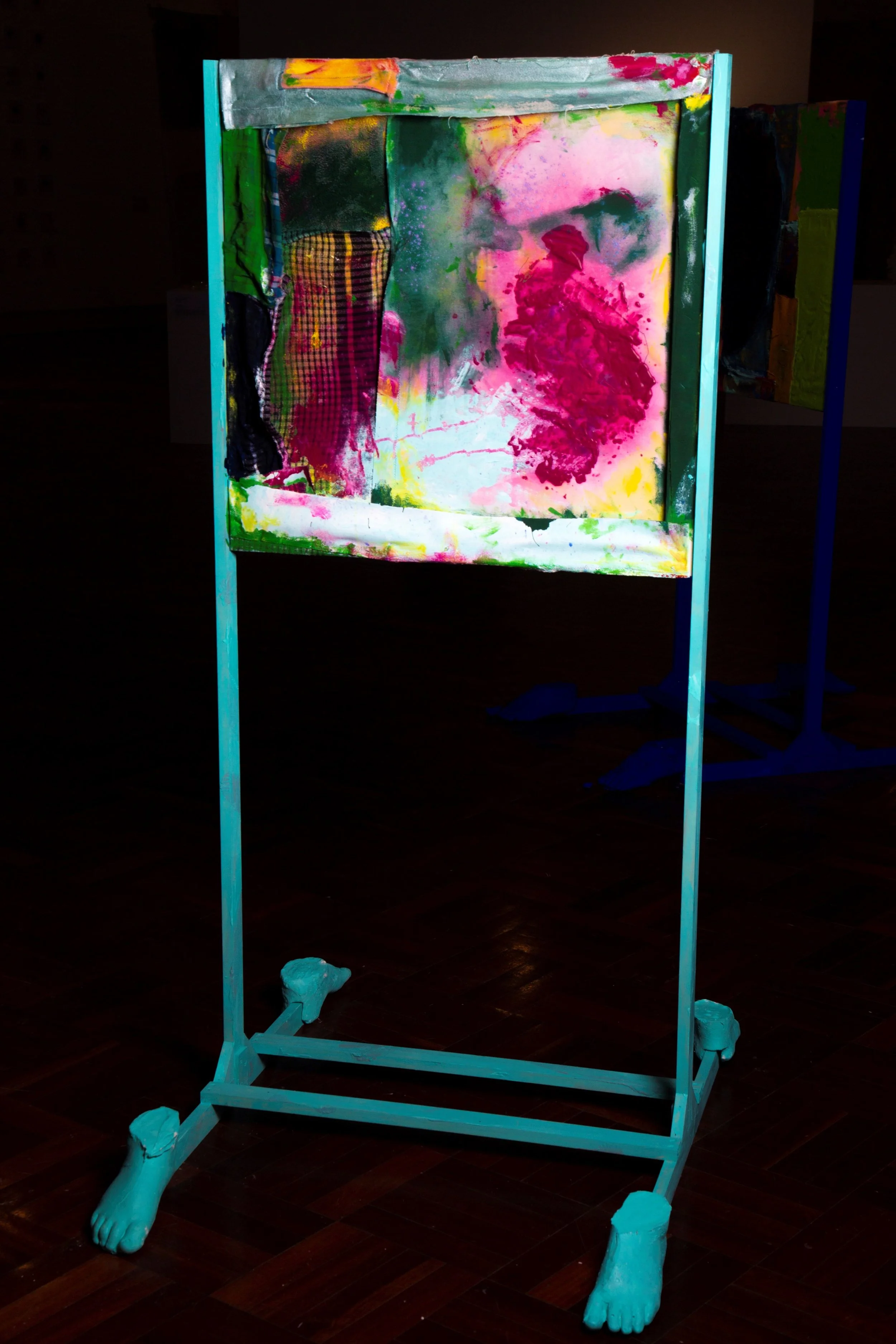 An art display featuring an abstract painting on a frame with painted feet at the base, set on a dark wooden floor.