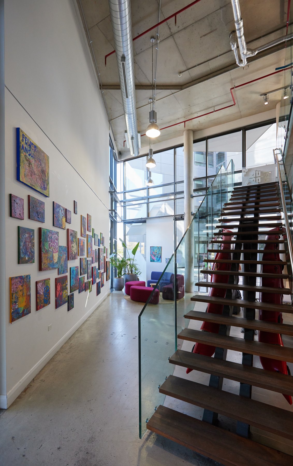 Modern art gallery interior with colorful paintings on the wall, a staircase with glass railing, and seating area with purple and blue chairs near large windows with natural light.