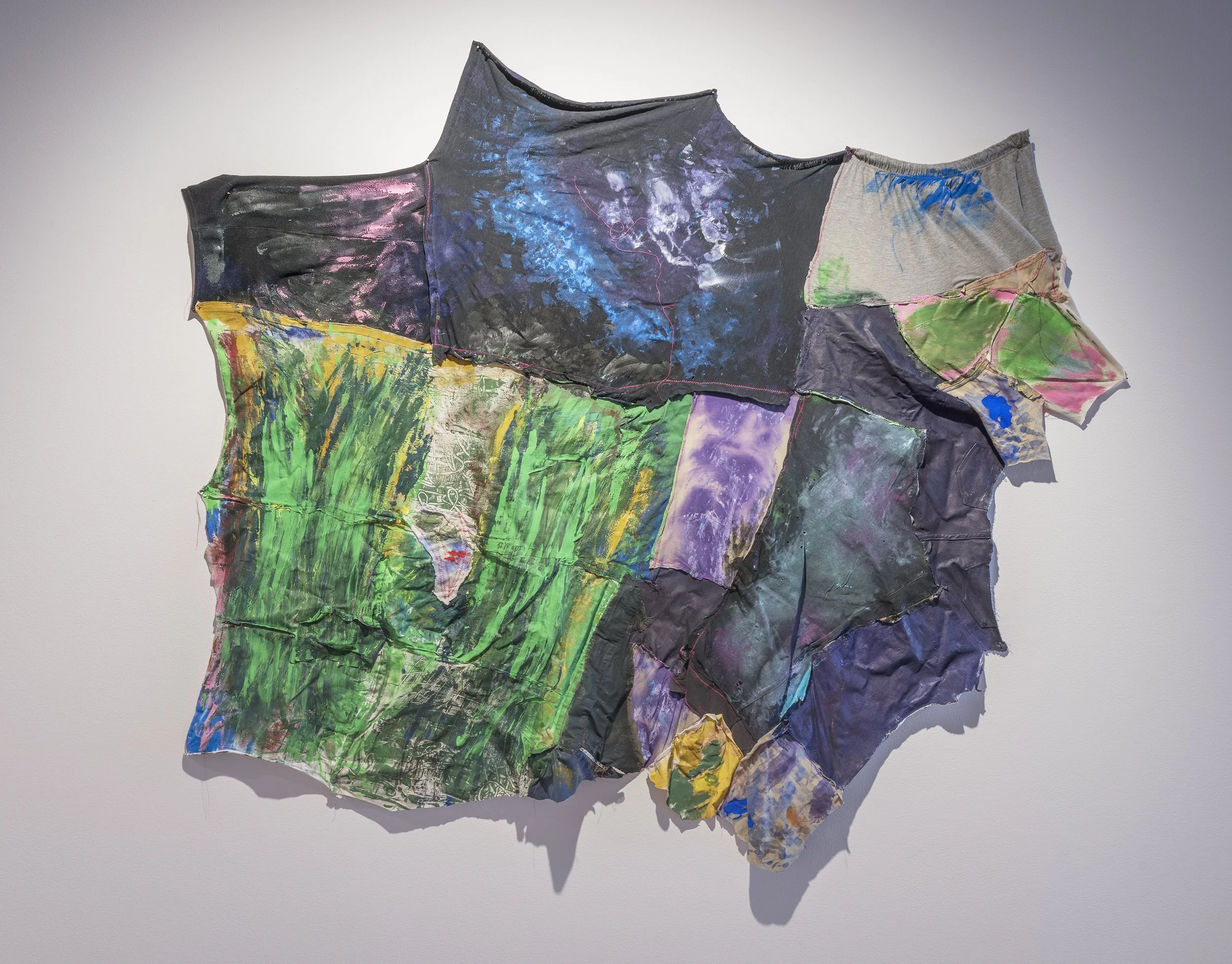 Mixed media abstract art piece made from fabric with colorful paint splashes and patterns, mounted on a white wall.