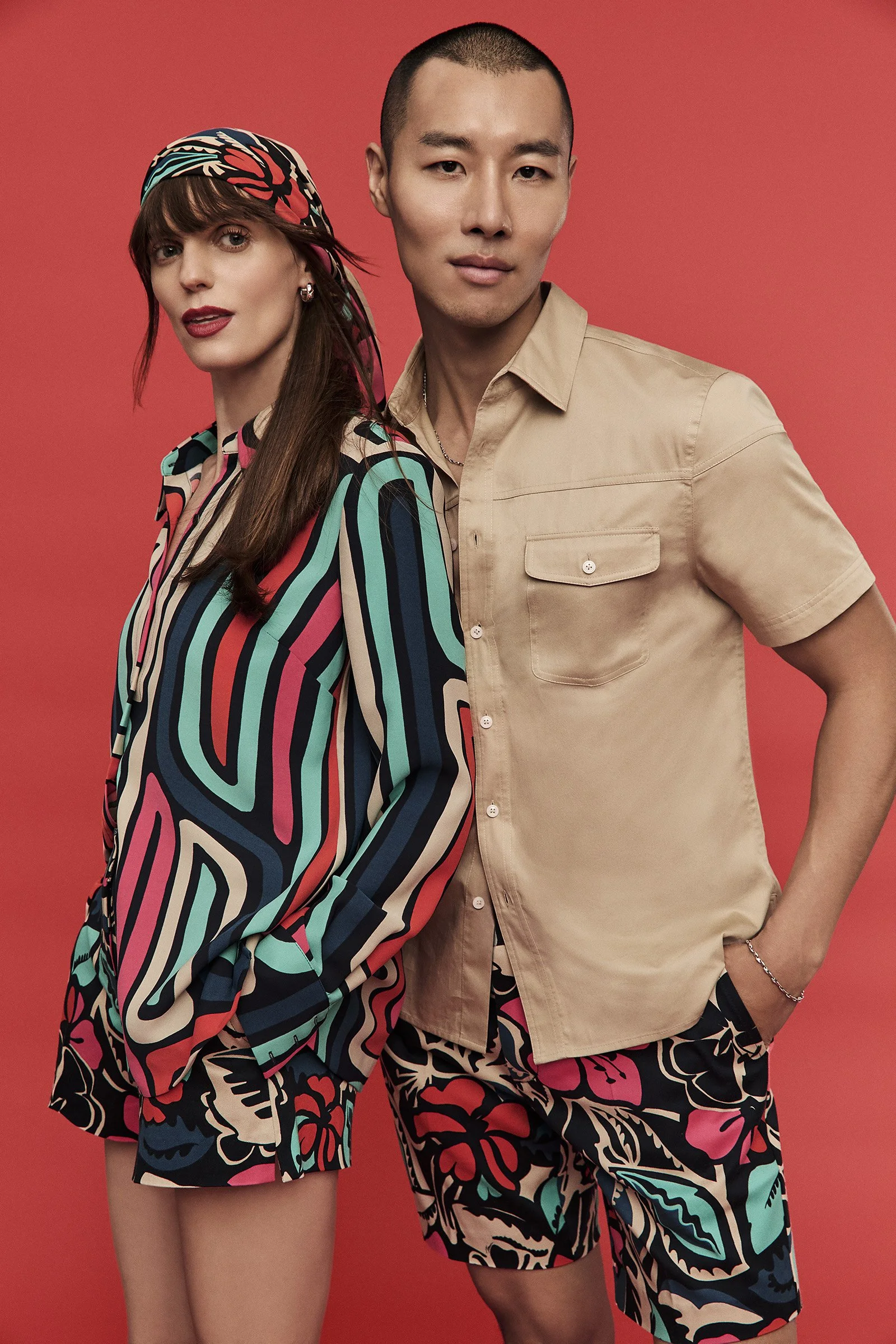 Two models, a woman with brown hair and a man with short black hair, standing against a red background, wearing colorful, patterned clothing.