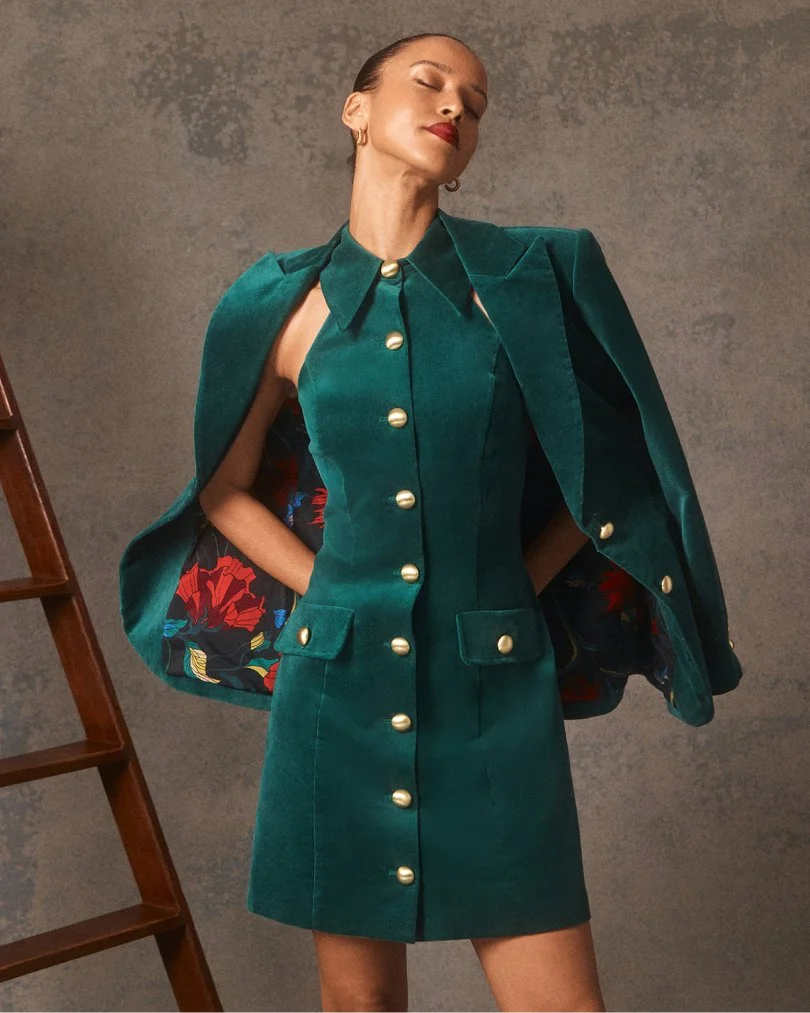 Woman in a green dress with pearl buttons and matching cape, standing with eyes closed against a textured background.