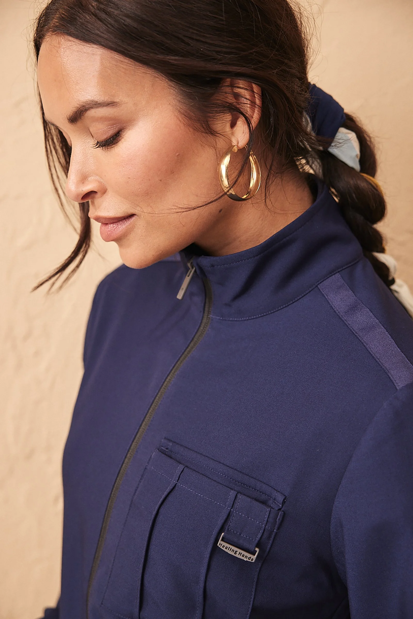 Close-up of a woman with her eyes closed, wearing gold hoop earrings, a navy jacket with a zipper, and a tag that reads 'Healing Hands', against a beige wall.