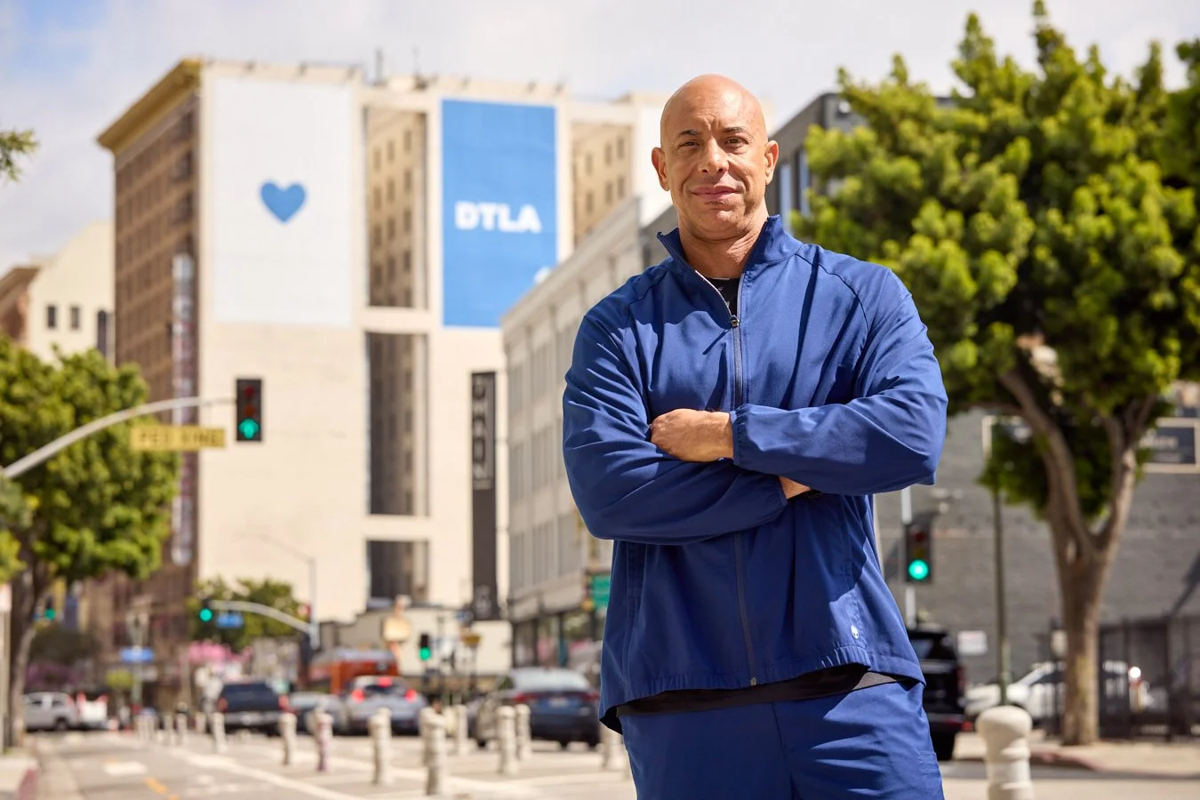 A man in a blue jacket crossing his arms standing on a city street with buildings, trees, and traffic lights in the background.