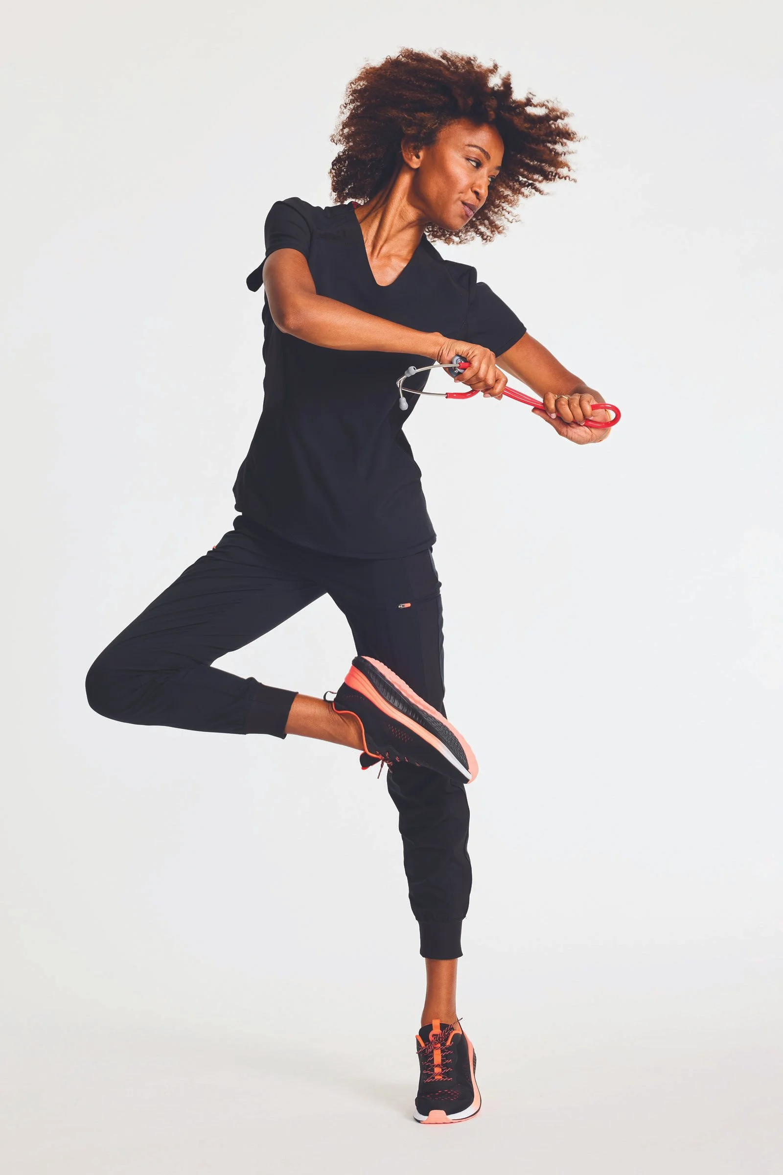 A woman with curly hair aerobics in a black workout outfit holding resistance bands, lifting one leg in a fitness pose against a plain background.