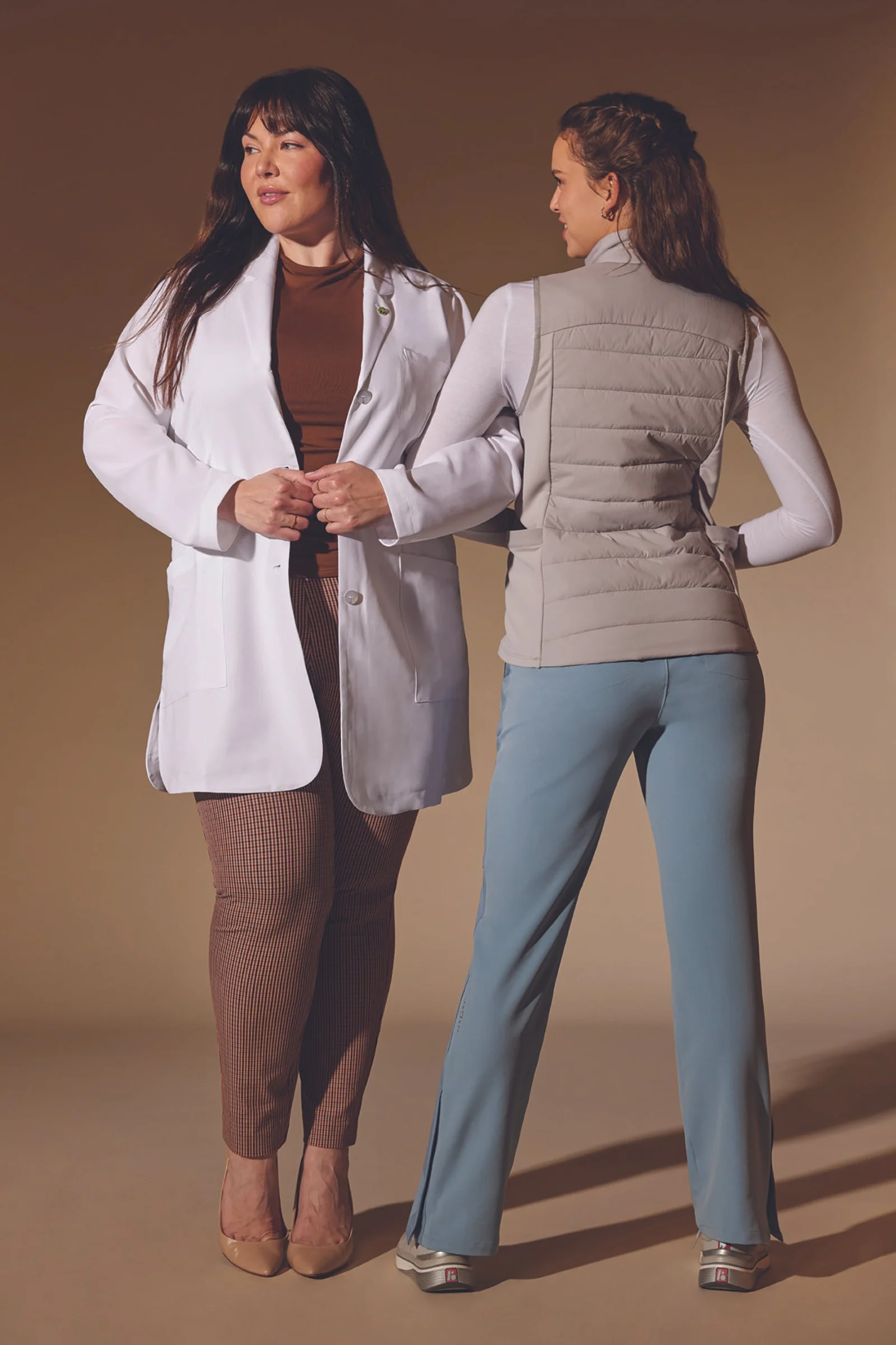 Two women stand facing each other, one in a white lab coat and the other in casual clothing, holding hands in a friendly gesture against a plain background.