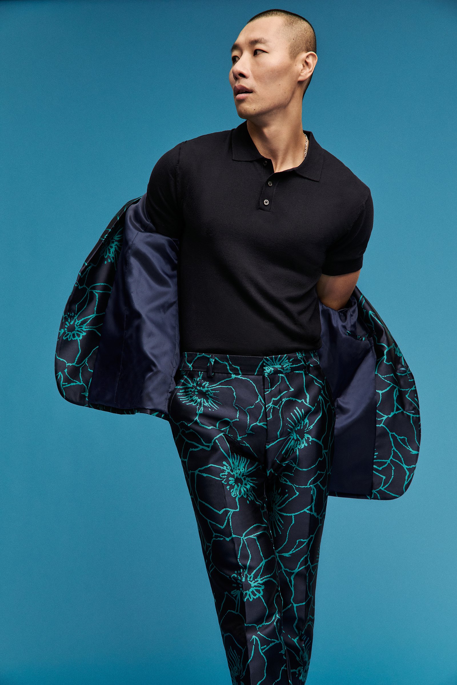 A man with a shaved head wearing a black polo shirt and patterned pants, holding a matching patterned jacket over his shoulder against a blue background.