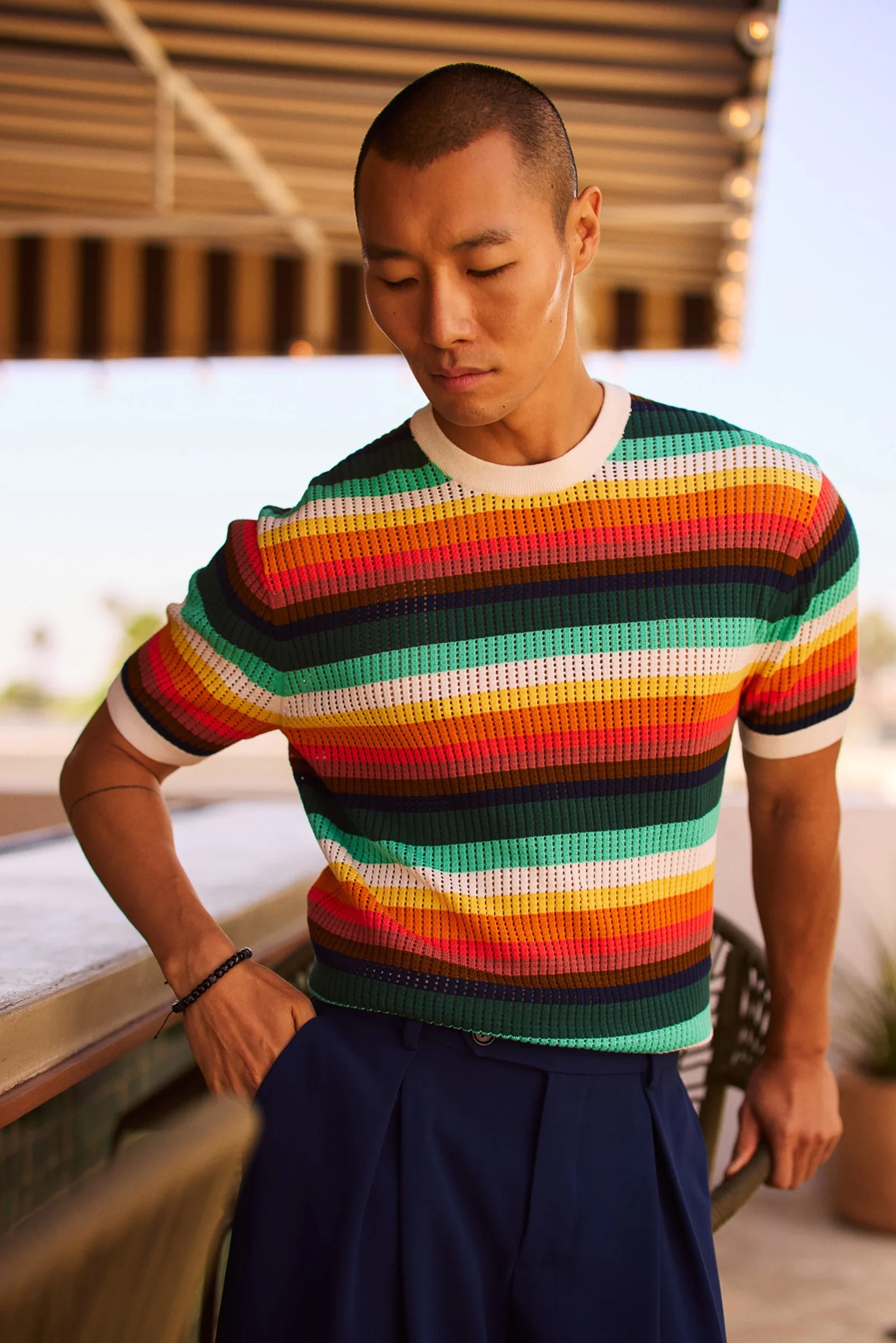 A man wearing a colorful, striped short-sleeve shirt and dark blue pants, standing on a patio with a wooden roof overhead, outdoors during daytime.