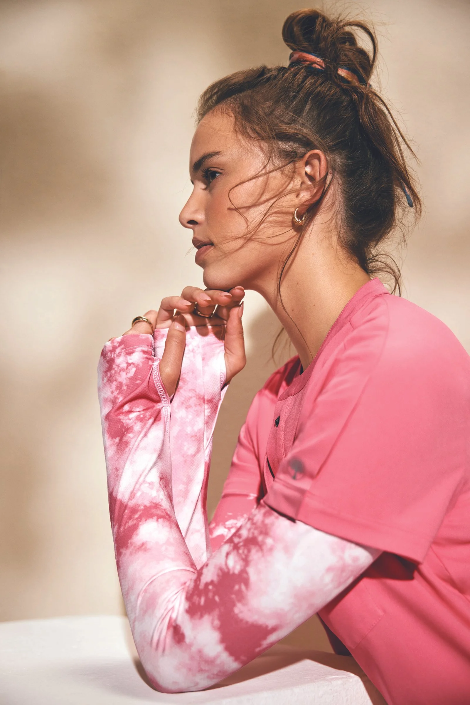 A young woman with wavy brown hair tied up in a bun with a pink scrunchie, wearing a pink sports shirt and a long sleeve tie-dye shirt, resting her chin on her hand.
