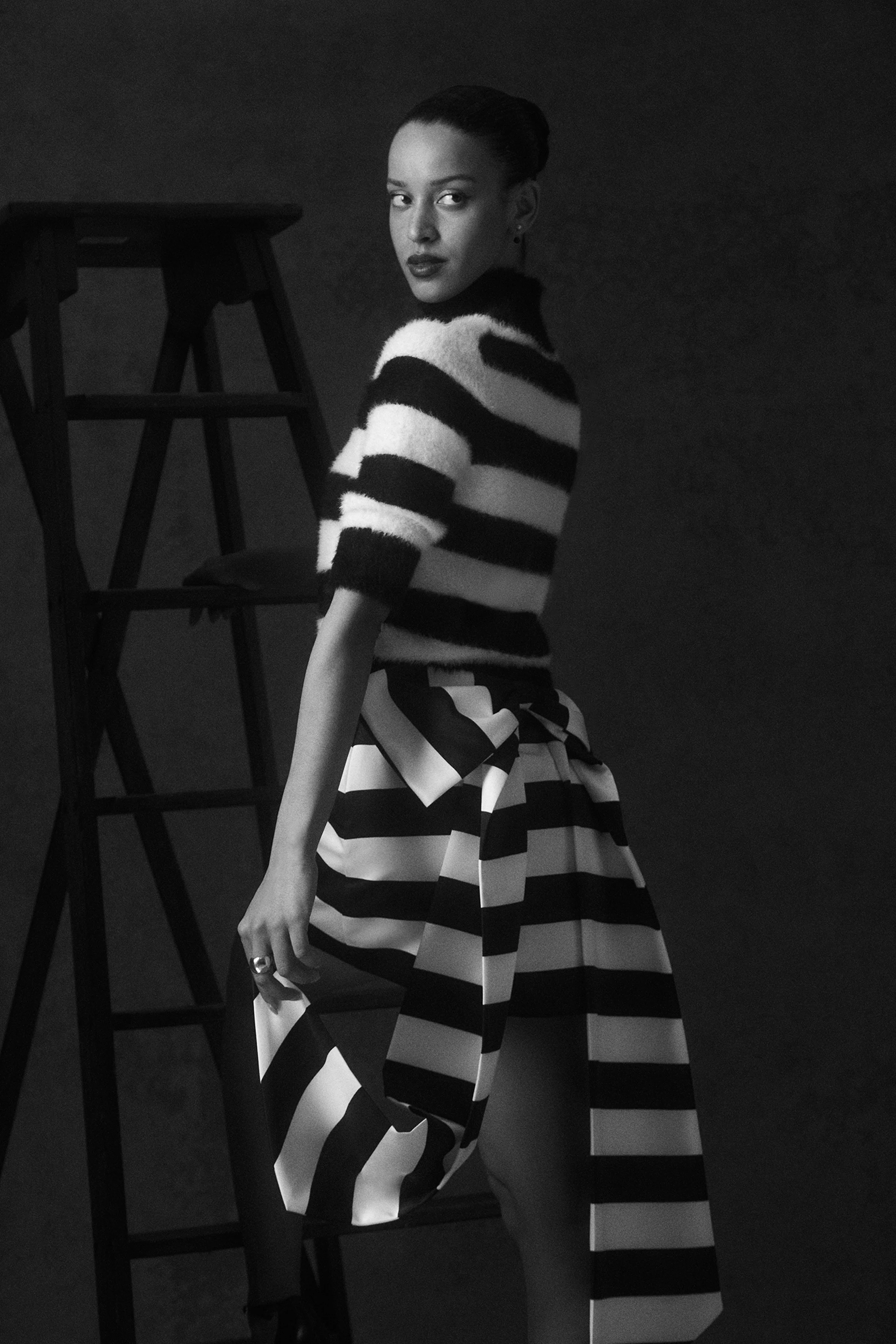 A black and white portrait of a woman with dark lipstick, wearing a black and white striped sweater and a matching striped skirt, standing next to a wooden ladder against a dark background.