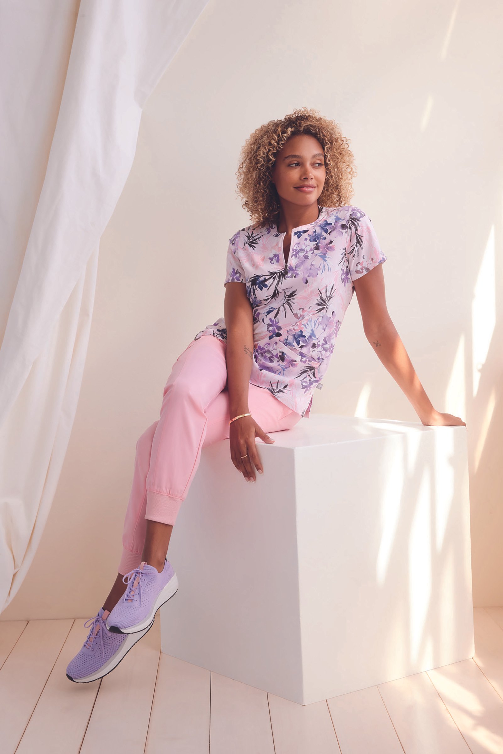 Woman sitting on a white block, wearing a floral purple and pink shirt, light pink pants, and purple sneakers, with a light-colored background and draped fabric on the side.