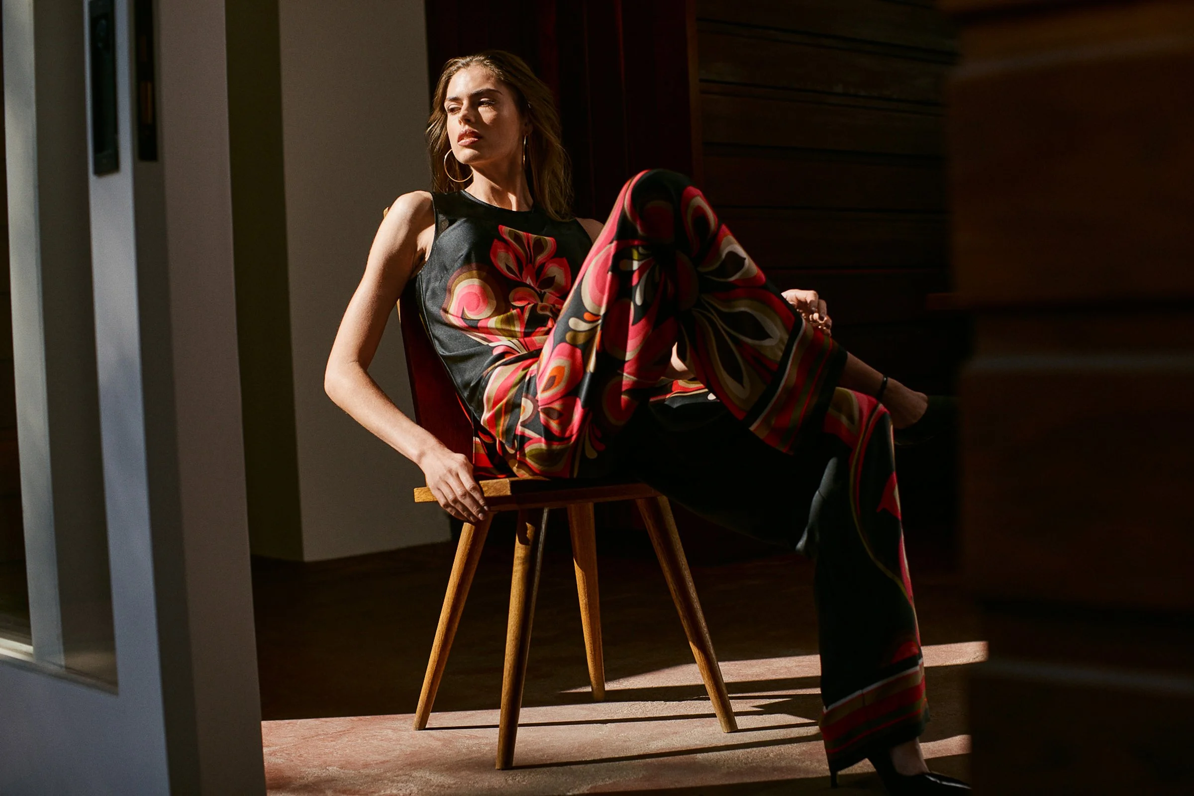 Fashion model sitting on a wooden chair in a dimly lit room, wearing a colorful silk outfit with a floral pattern.