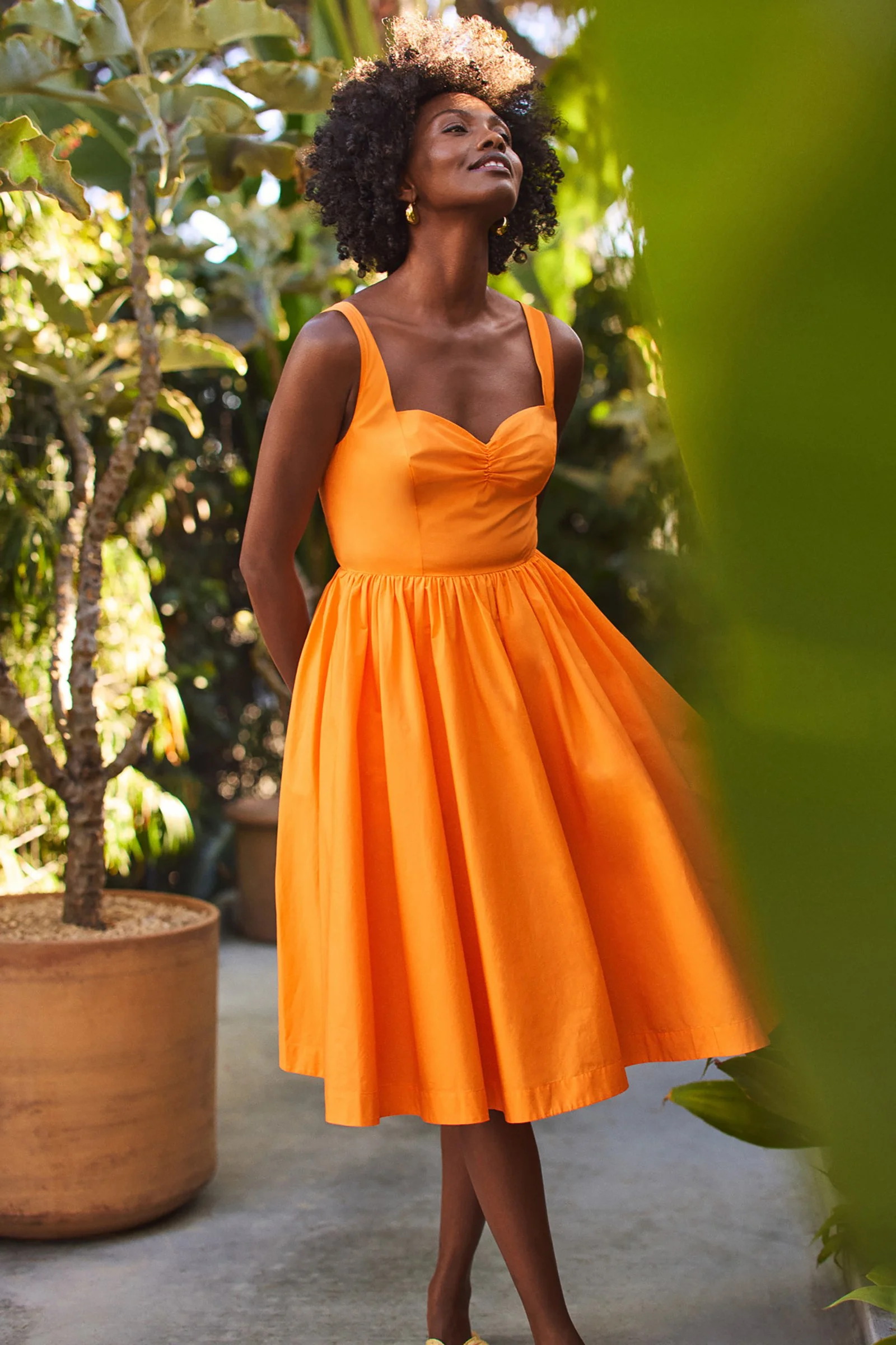 A woman in an orange dress standing outdoors among green plants and trees, smiling with her eyes closed.