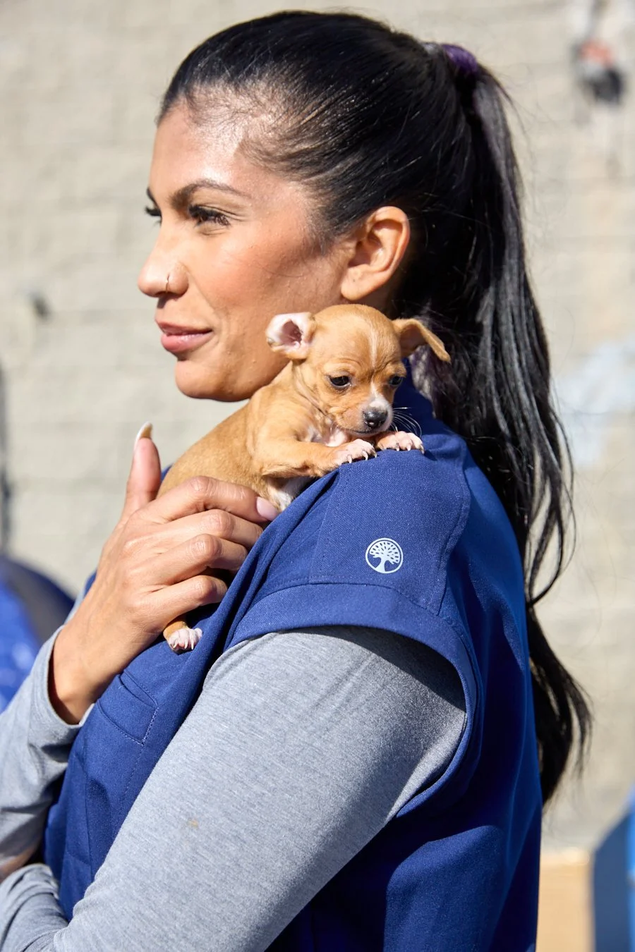 A woman with dark hair in a ponytail holding a small tan puppy on her shoulder.