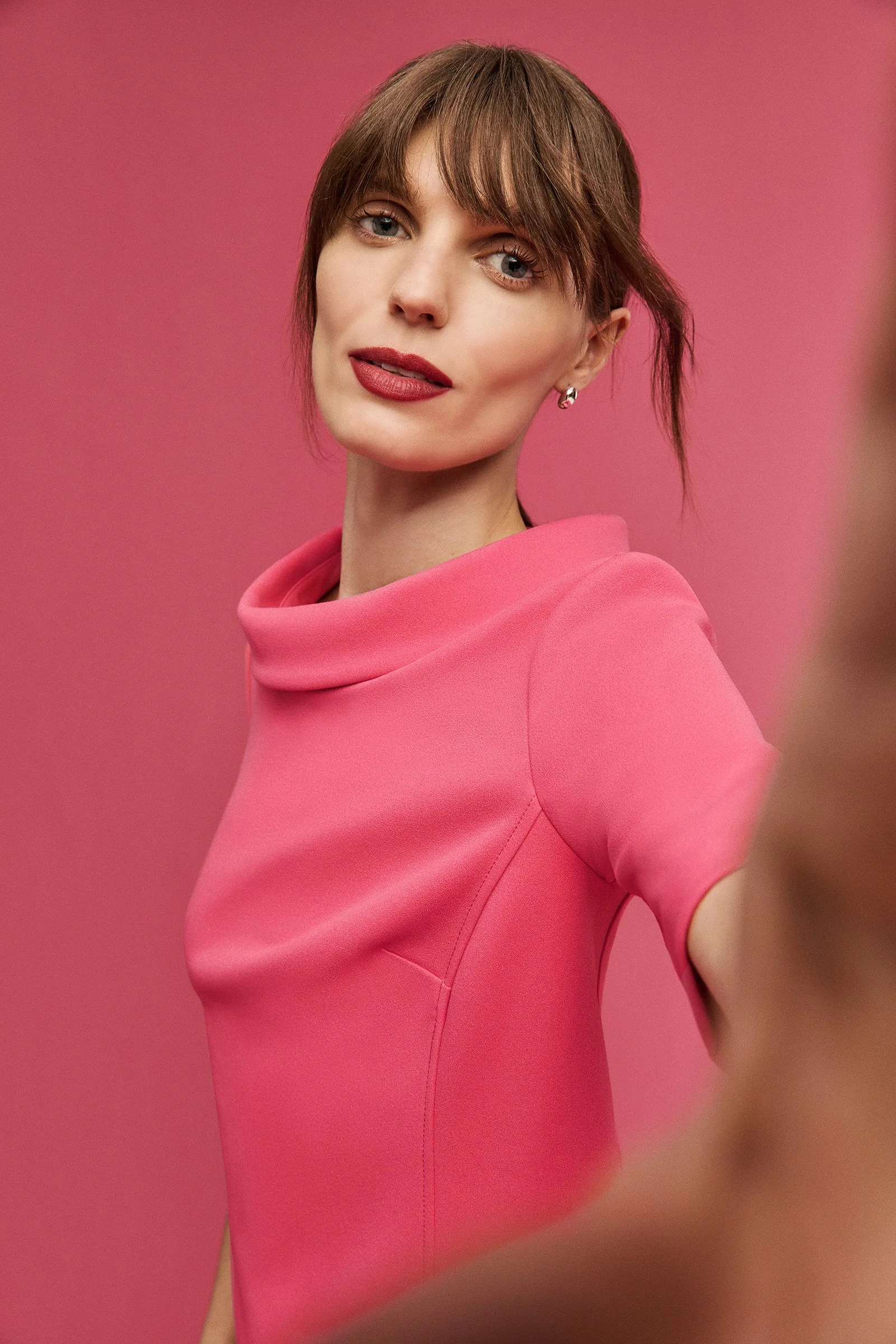 A woman with short brown hair and blue eyes wearing a pink top, taking a selfie against a pink background.