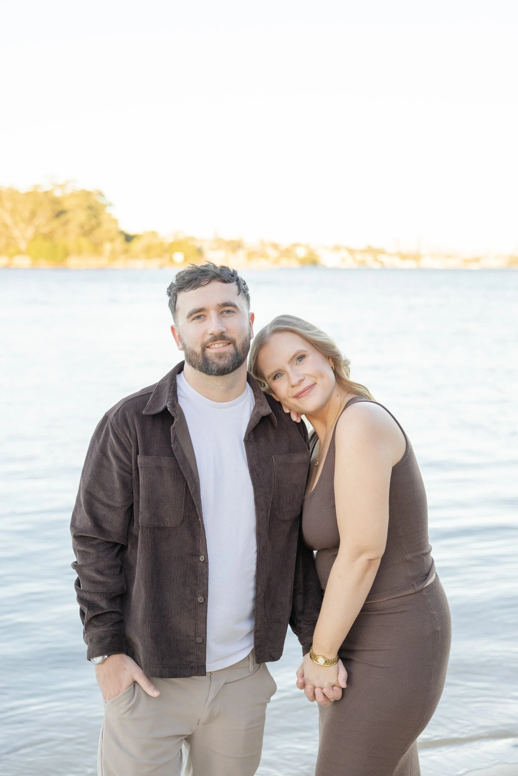 A man with a beard and short dark hair, wearing a brown jacket and white t-shirt, standing next to a woman with blonde hair, wearing a brown dress, by the water with a distant shoreline and trees in the background. They are holding hands and smiling.