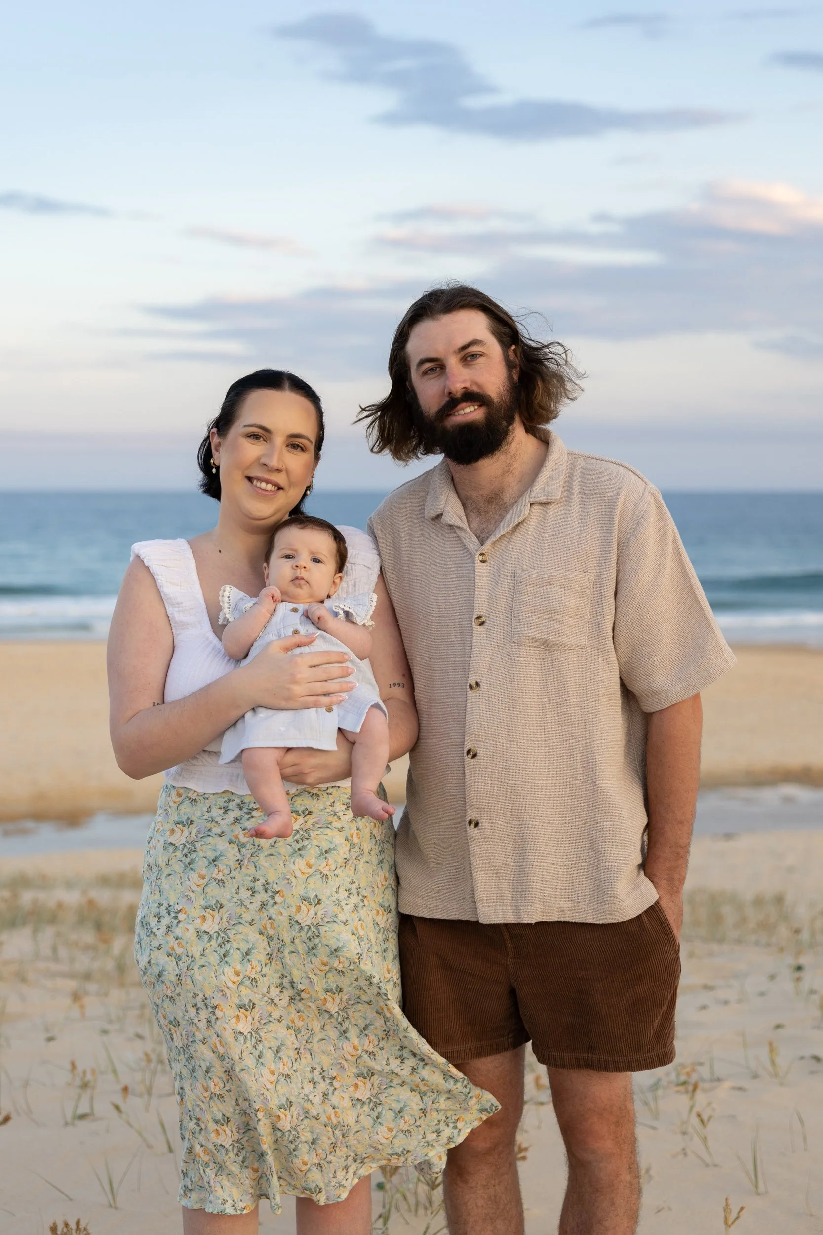 A family of three on a beach during sunset, with the mother holding a baby girl, and the father standing beside them, all smiling.