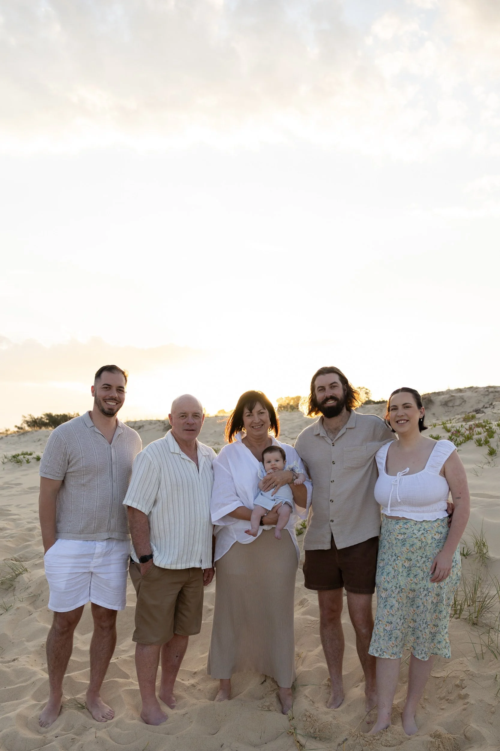 A group of five adults and one baby standing on a sandy beach during sunset, smiling at the camera.