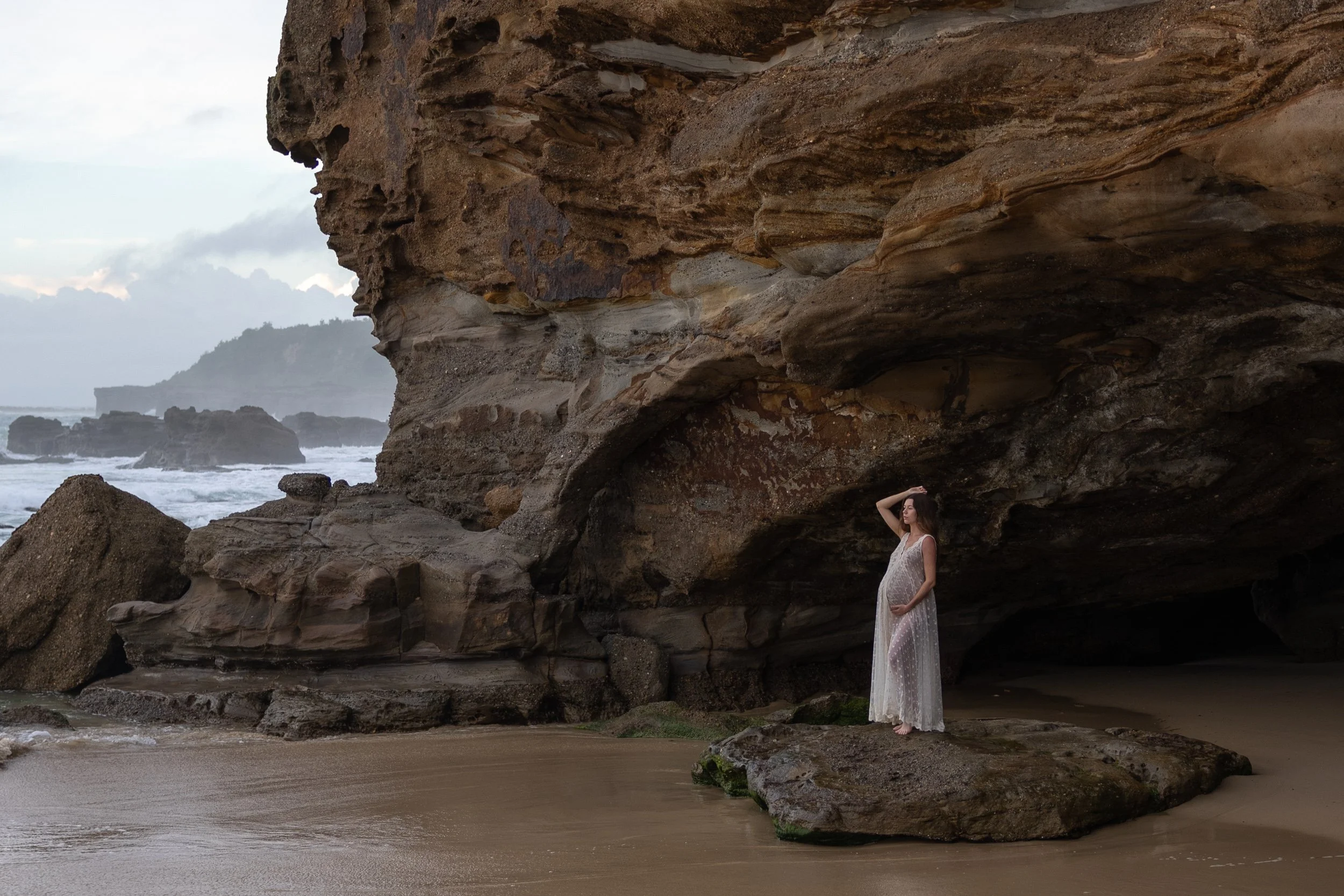 A woman in a white, semi-transparent dress with long hair standing on a rocky beach beneath large rock formations near the ocean with waves and cloudy sky in the background.