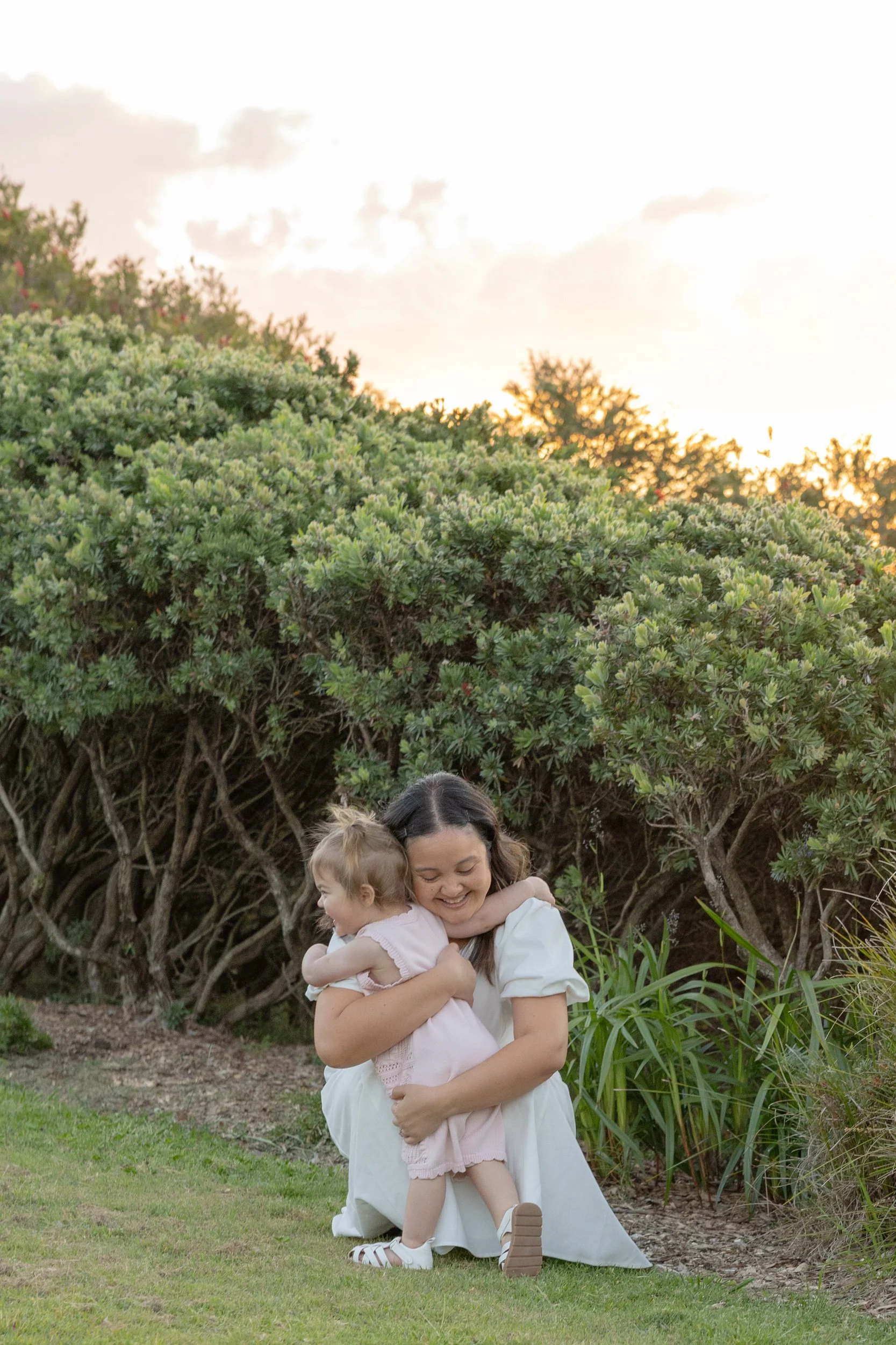 A woman hugging her toddler outdoors near green bushes and grass with the sun setting in the background.