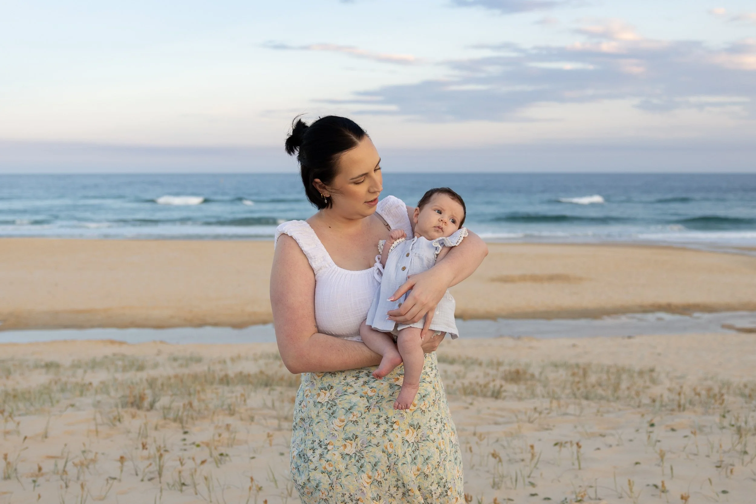 A woman holding a baby on a sandy beach with the ocean in the background at sunset.
