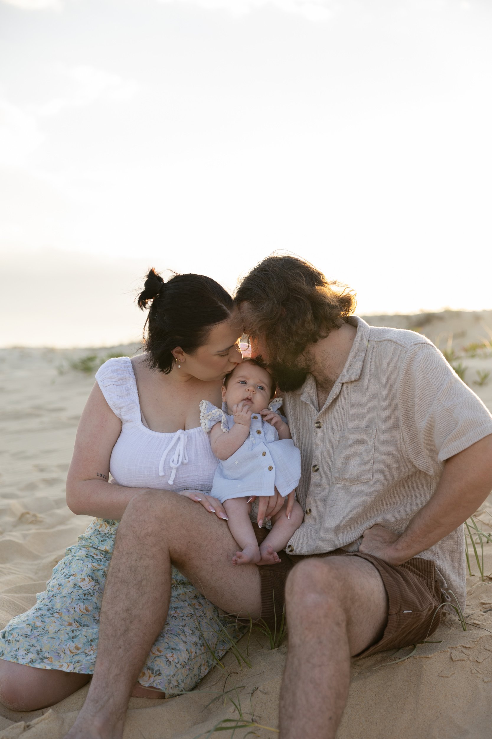 A family of three sitting on the beach, with the mother and father kissing their baby on the forehead, during sunset.