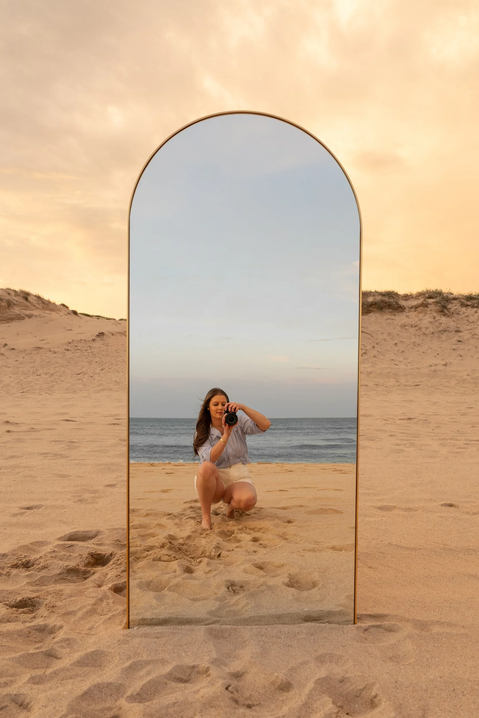 A woman taking a mirror selfie on a sandy beach at sunset, with the ocean in the background and sky with soft orange clouds.