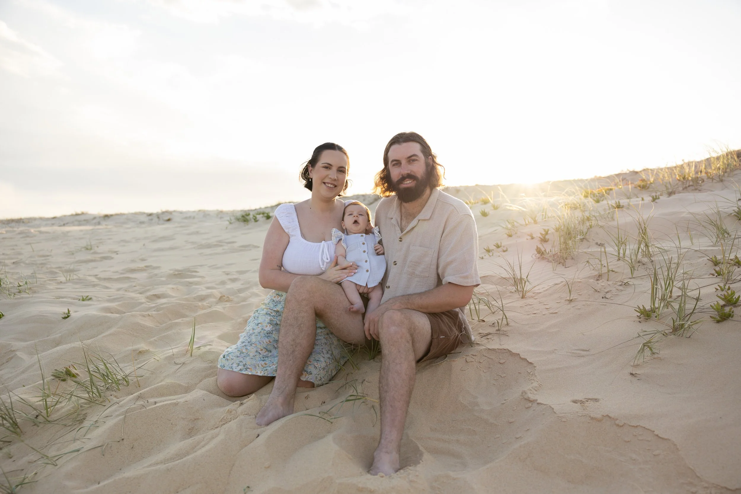 A family of three sitting on sandy beach with beach grass, smiling at the camera during sunset.