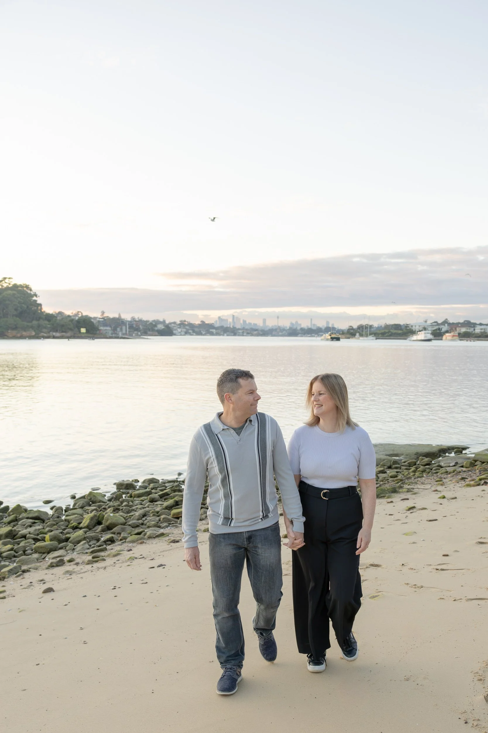 A couple walking hand in hand on a beach with rocks, water, and city skyline in the background during sunrise.