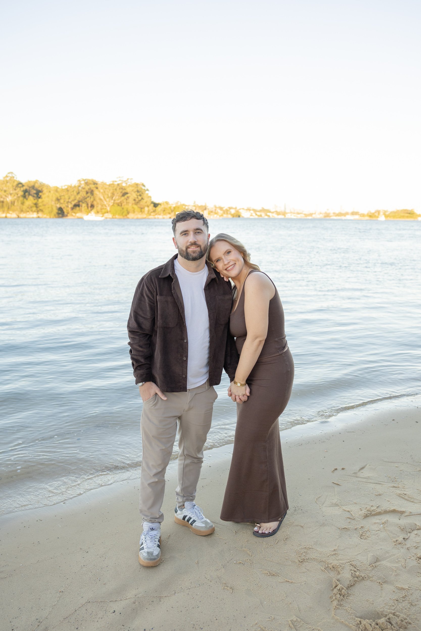A couple standing close together on a beach near the water, smiling at the camera with land and trees in the background. The man has dark hair and a beard, wearing a dark jacket and beige pants. The woman is wearing a brown dress and sandals.