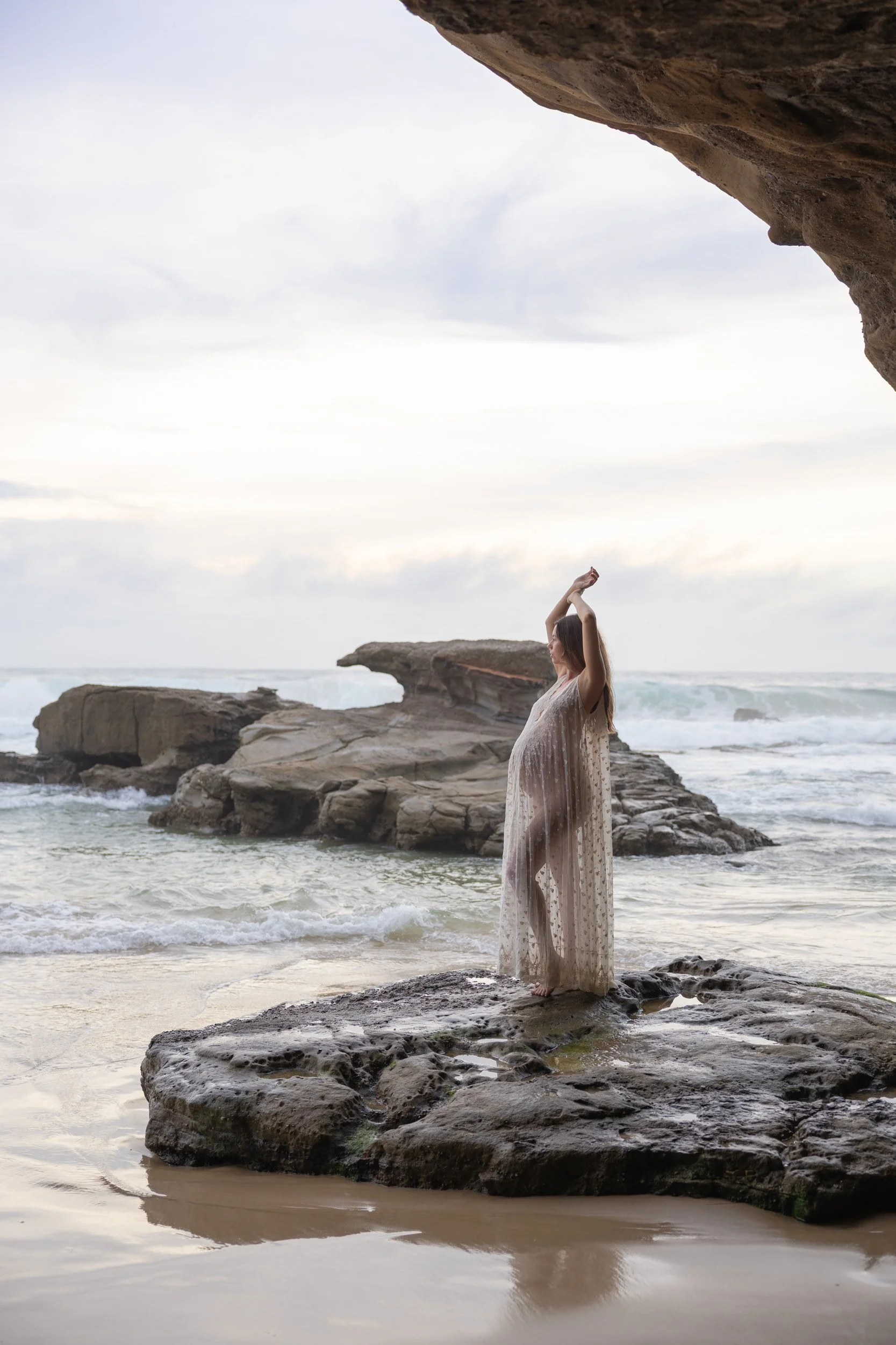 A woman in a long, semi-transparent dress standing on rocks at a beach, with large rock formations over her head and waves in the background.