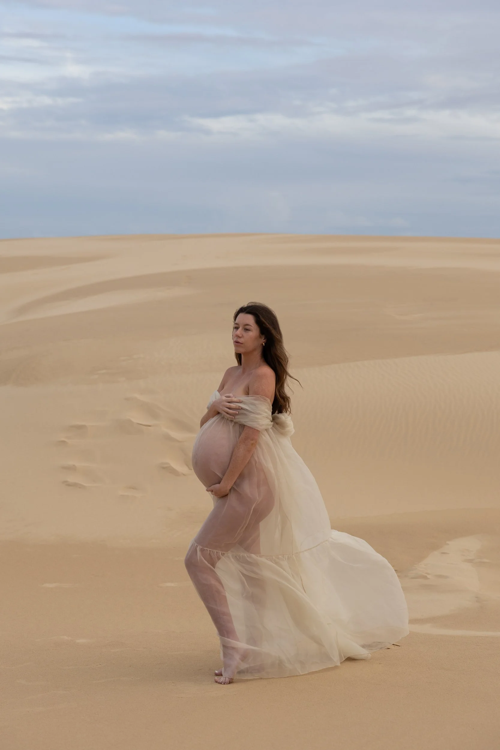 A pregnant woman in a flowing, semi-transparent white dress standing barefoot on sand dunes, holding her belly with both hands, under a partly cloudy sky.