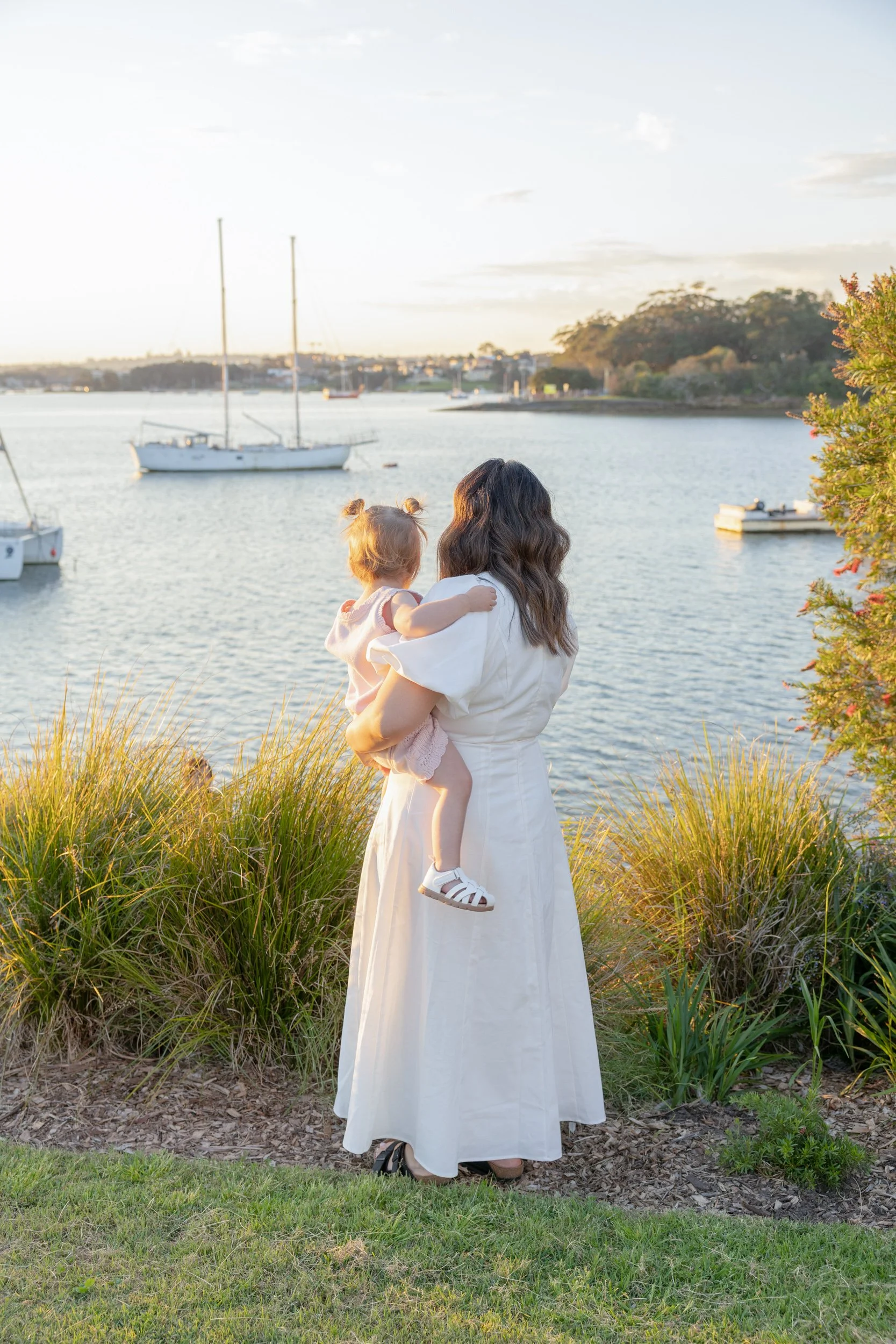 A woman holding her daughter in her arms, standing outdoors near a river with boats and sailboats, trees, and hills in the background during sunset.