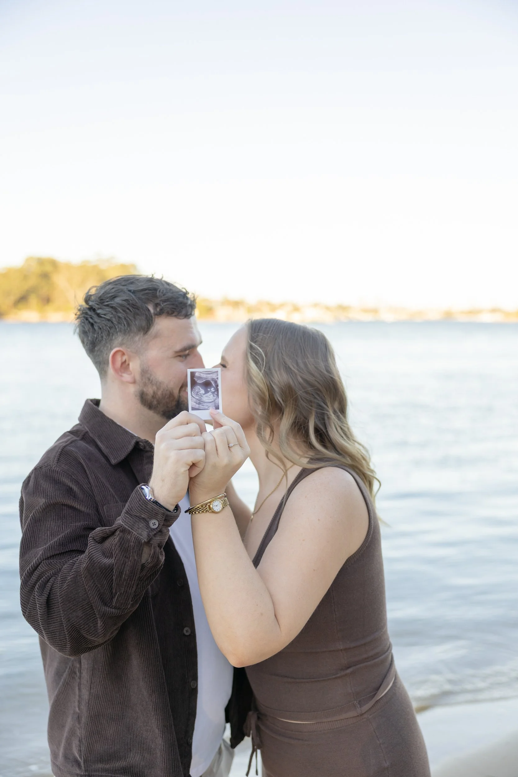 A couple at the beach holding an ultrasound picture, nose to nose, with the water and sky in the background.