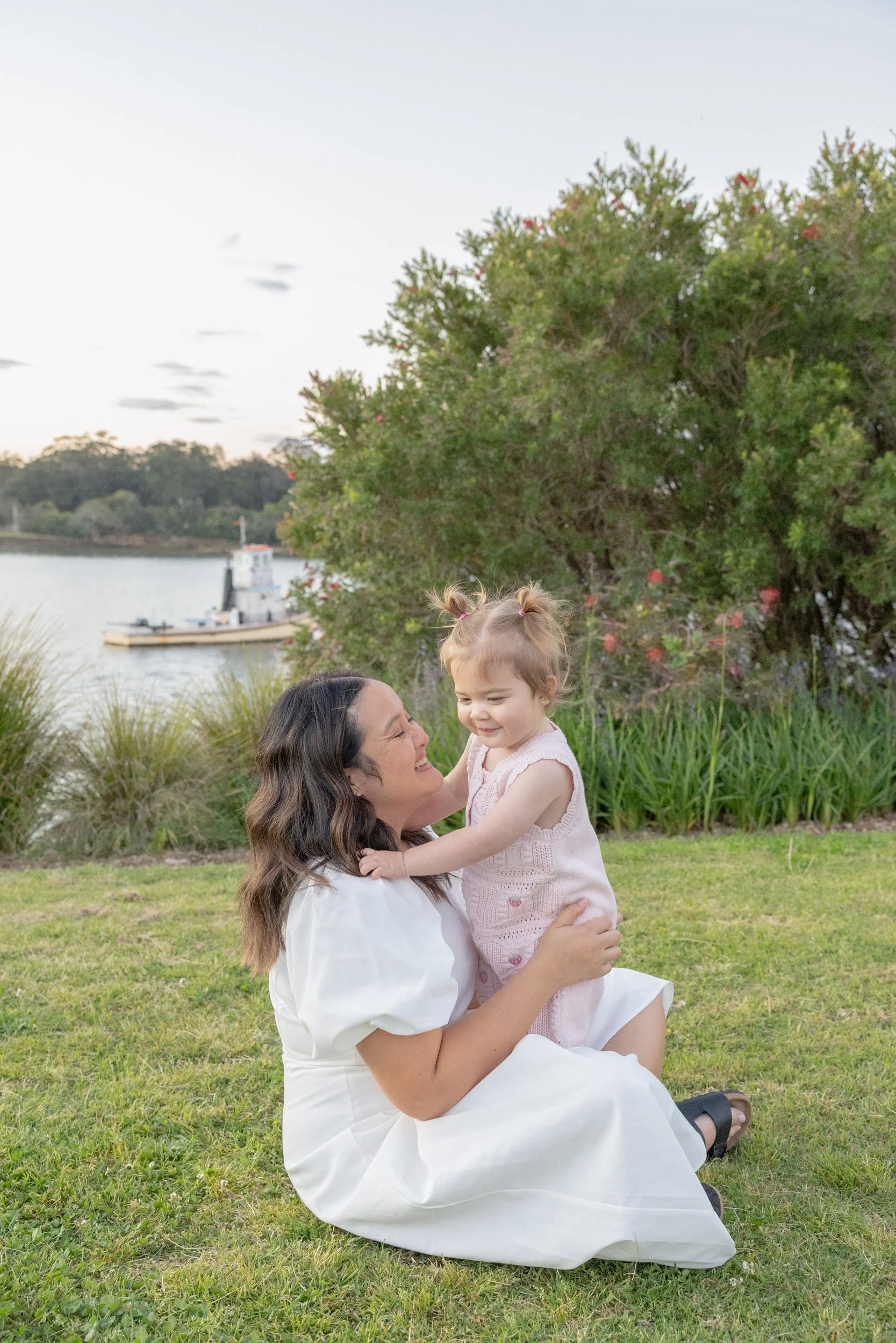 A woman with long brown hair sitting on grass, holding a young girl with light brown hair and pigtails, both smiling and enjoying outdoors near water and trees at sunset.