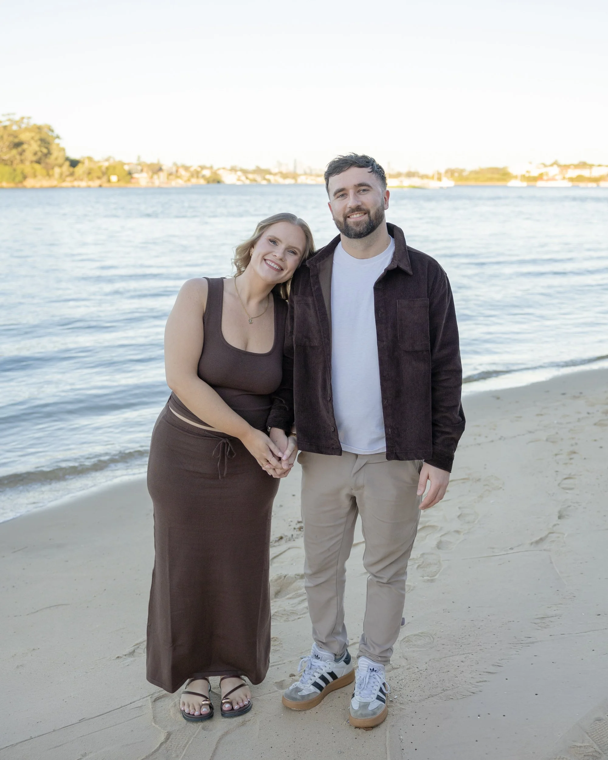 A man and woman standing hand in hand on a sandy beach with water and trees in the background. The woman is smiling with her head tilted, and the man is smiling with beard and short hair, wearing sneakers. The woman is wearing a sleeveless brown dress and sandals, and the man is in beige pants and white sneakers.