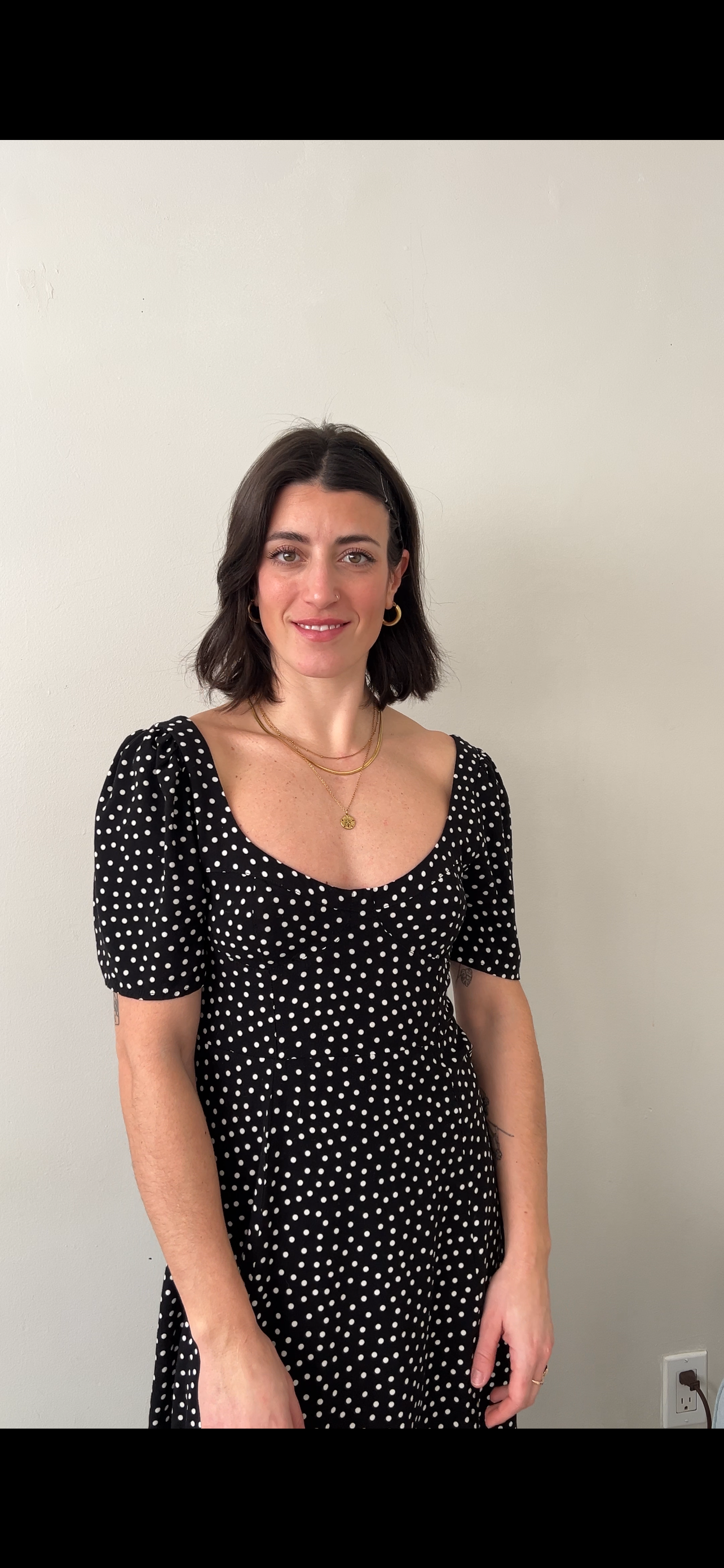 A woman with dark hair, wearing a black dress with white polka dots, gold jewelry, standing in front of a plain light-colored wall.
