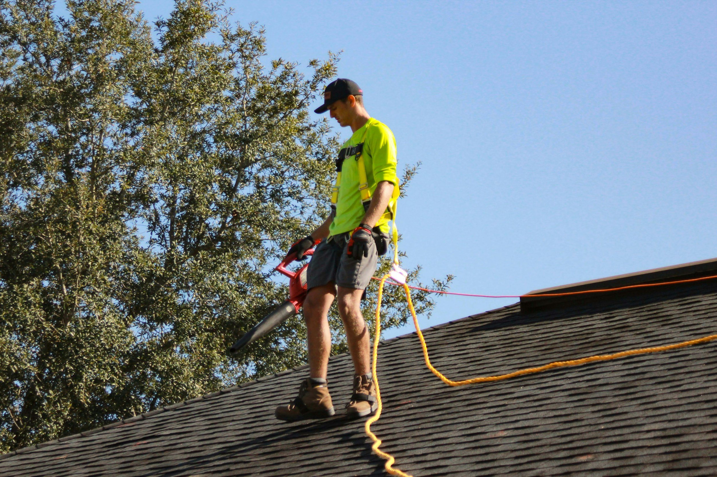A man standing on a rooftop, wearing a high-visibility shirt, shorts, gloves, and a cap, while holding a leaf blower, with a tree in the background.