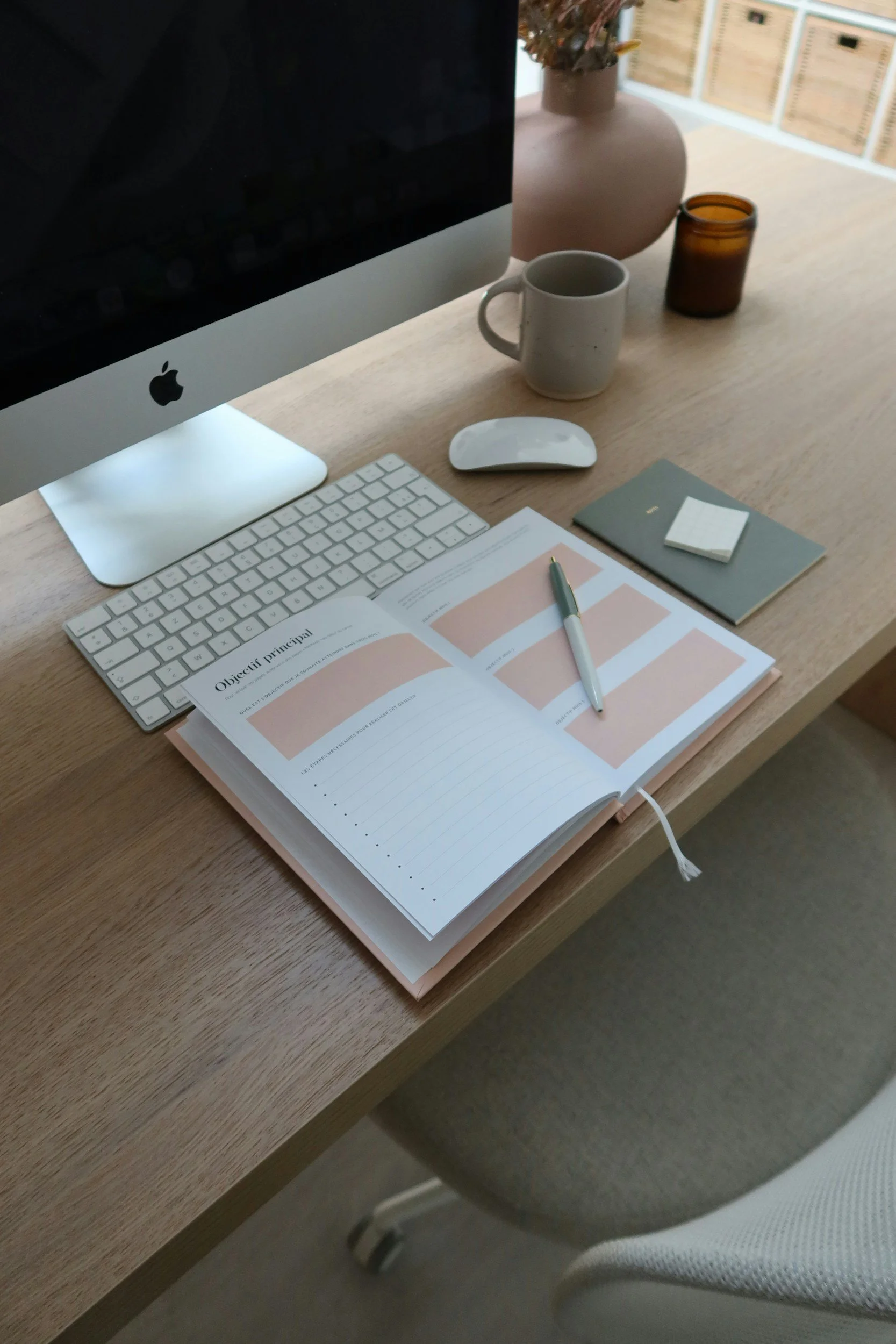 A tidy wooden desk with an iMac computer, white keyboard, mouse, open planner, pen, small notepad, mug, brown glass container, and a pink vase with flowers. There is a chair partially visible at the bottom right.