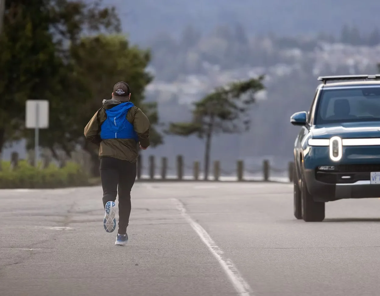 A person jogging on an empty road near a blue parked vehicle, with trees and mountains in the background.