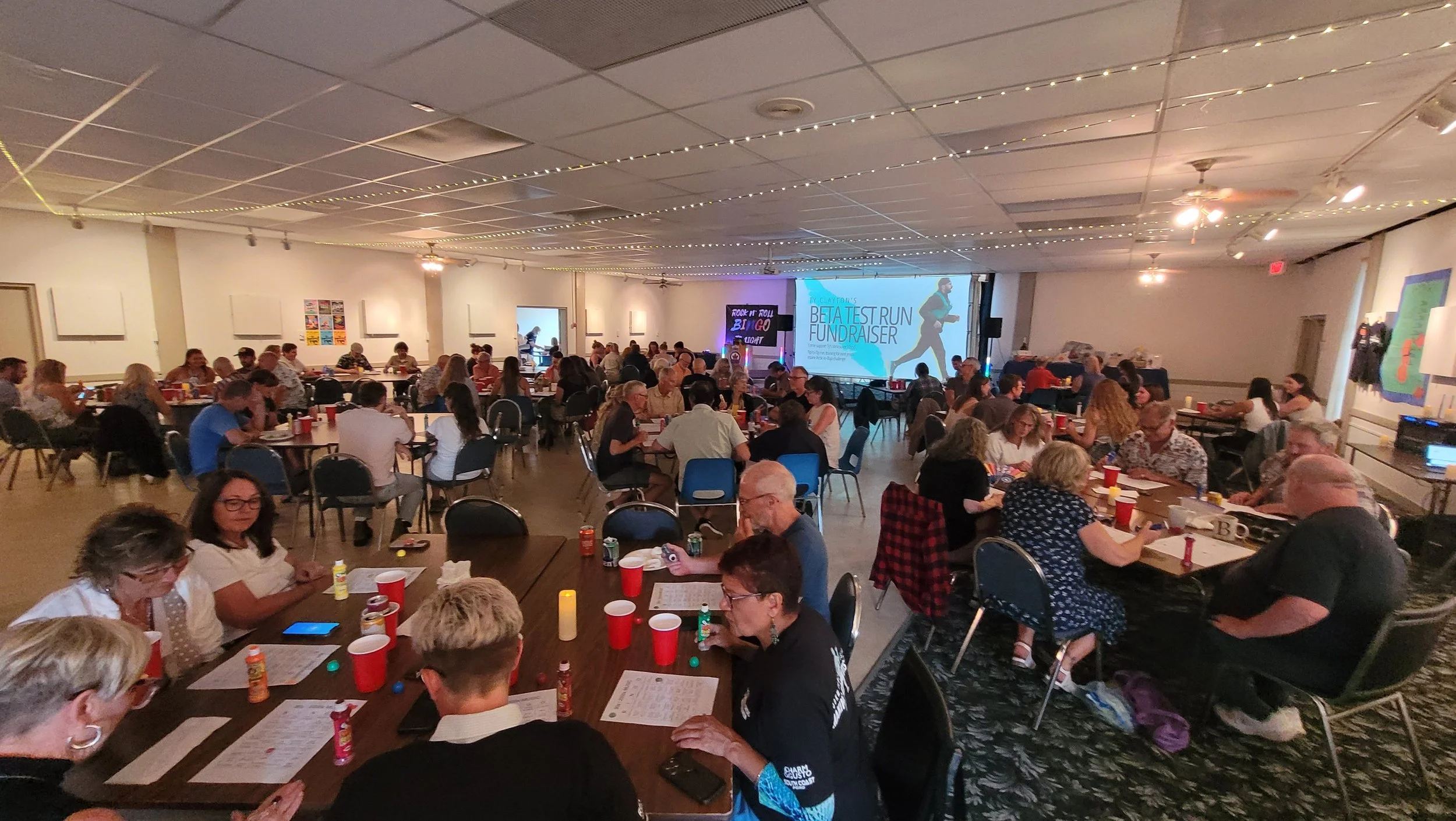 A large indoor gathering or fundraiser with multiple tables of people seated, some looking at papers, and a stage at the front displaying a projection screen with the screen text reading 'BET TEST RUN FUNDRAISER.' The room is decorated with string lights on the ceiling, and some artwork and signs on the walls.